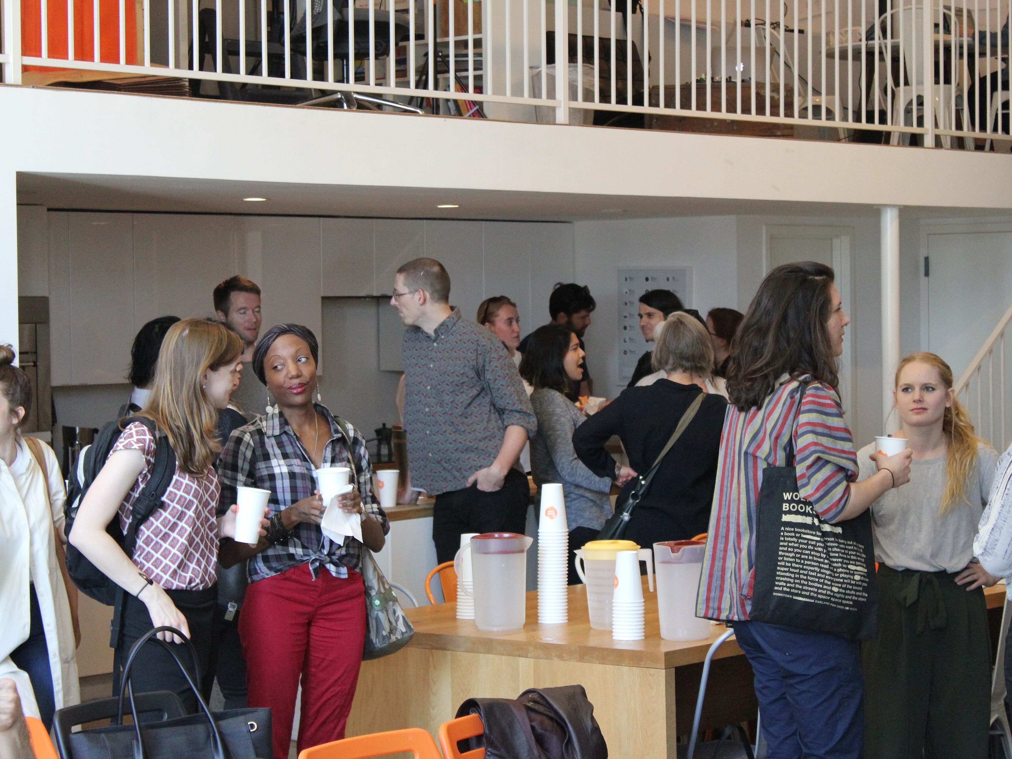 A group of people socializing and drinking from white cups in a brightly lit room with white walls. Some are standing while others are seated. There’s a countertop in the background with more cups, and a staircase and a mezzanine are visible.