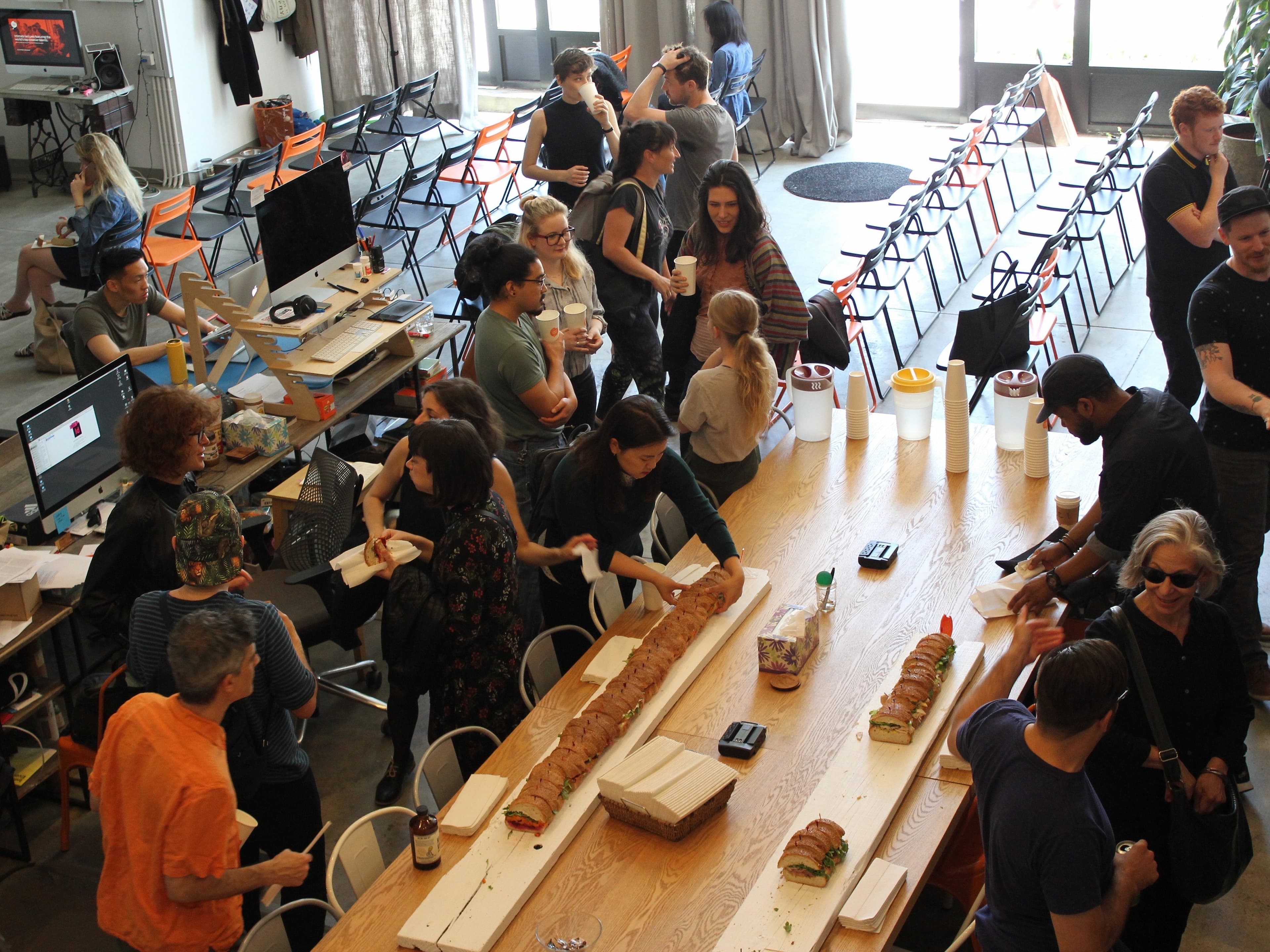 An overhead view of a lively gathering in a spacious room with large windows. People are socializing and eating sandwiches from a long table filled with food. Chairs are set up in rows, and desks with computers are visible in the background.