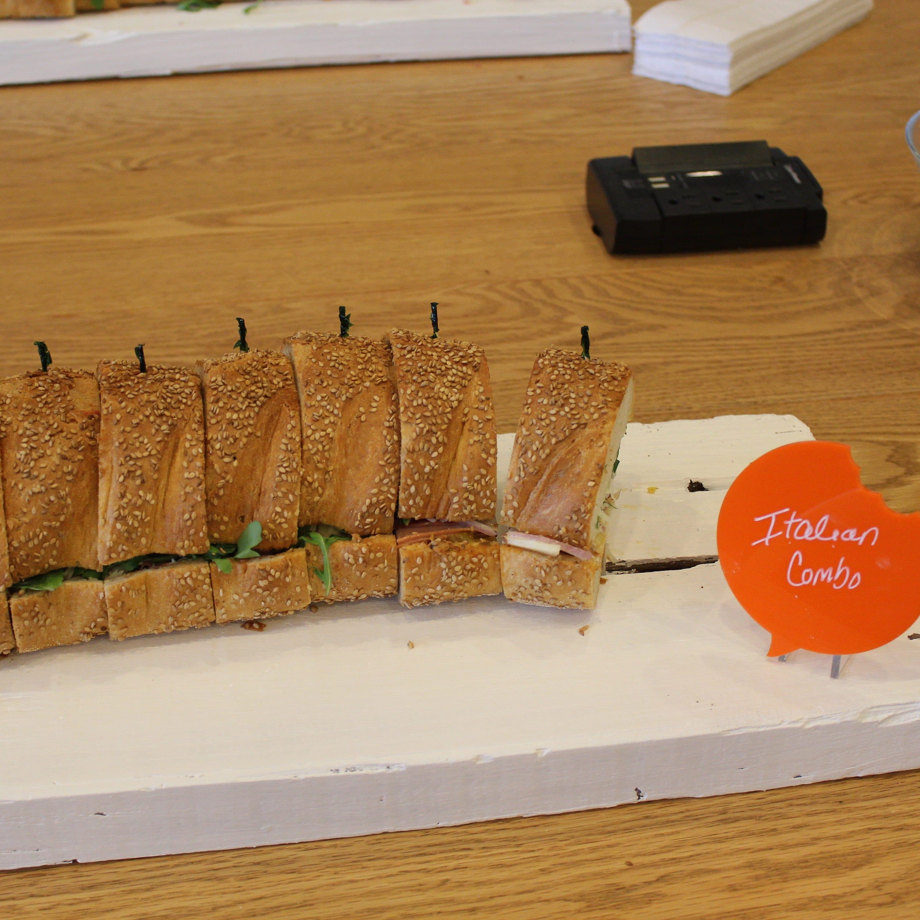 A tray of Italian combo sub sandwiches cut into smaller pieces is displayed on a wooden table. The sandwiches, with visible lettuce and deli meats, are placed on a white wooden board. A red speech bubble-shaped label reads "Italian Combo.