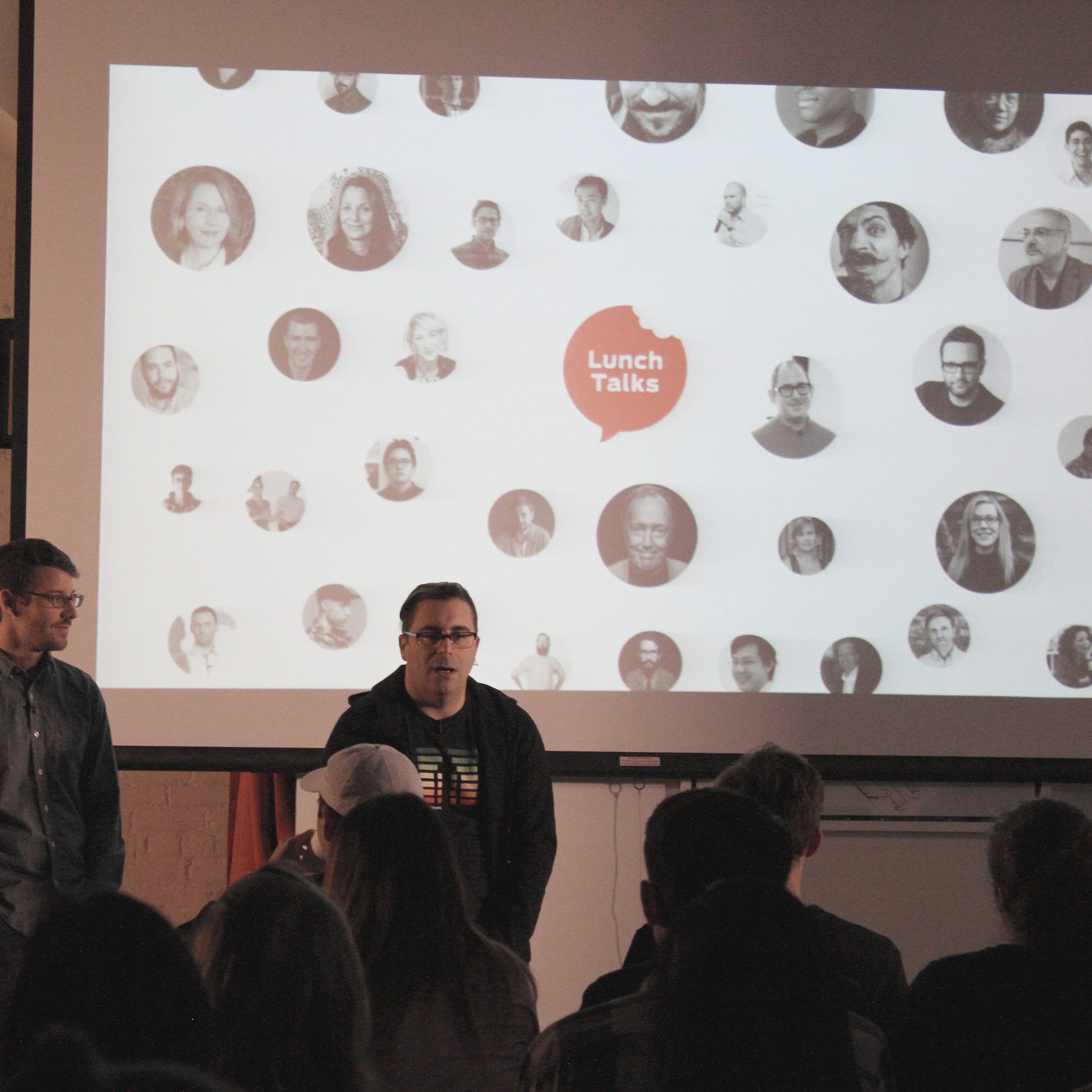 Two presenters stand in front of a projector screen displaying a collage of black-and-white headshots and a red speech bubble with the text "Lunch Talks." An audience is seated facing the presenters and the screen in a dimly lit room with framed pictures on the walls.