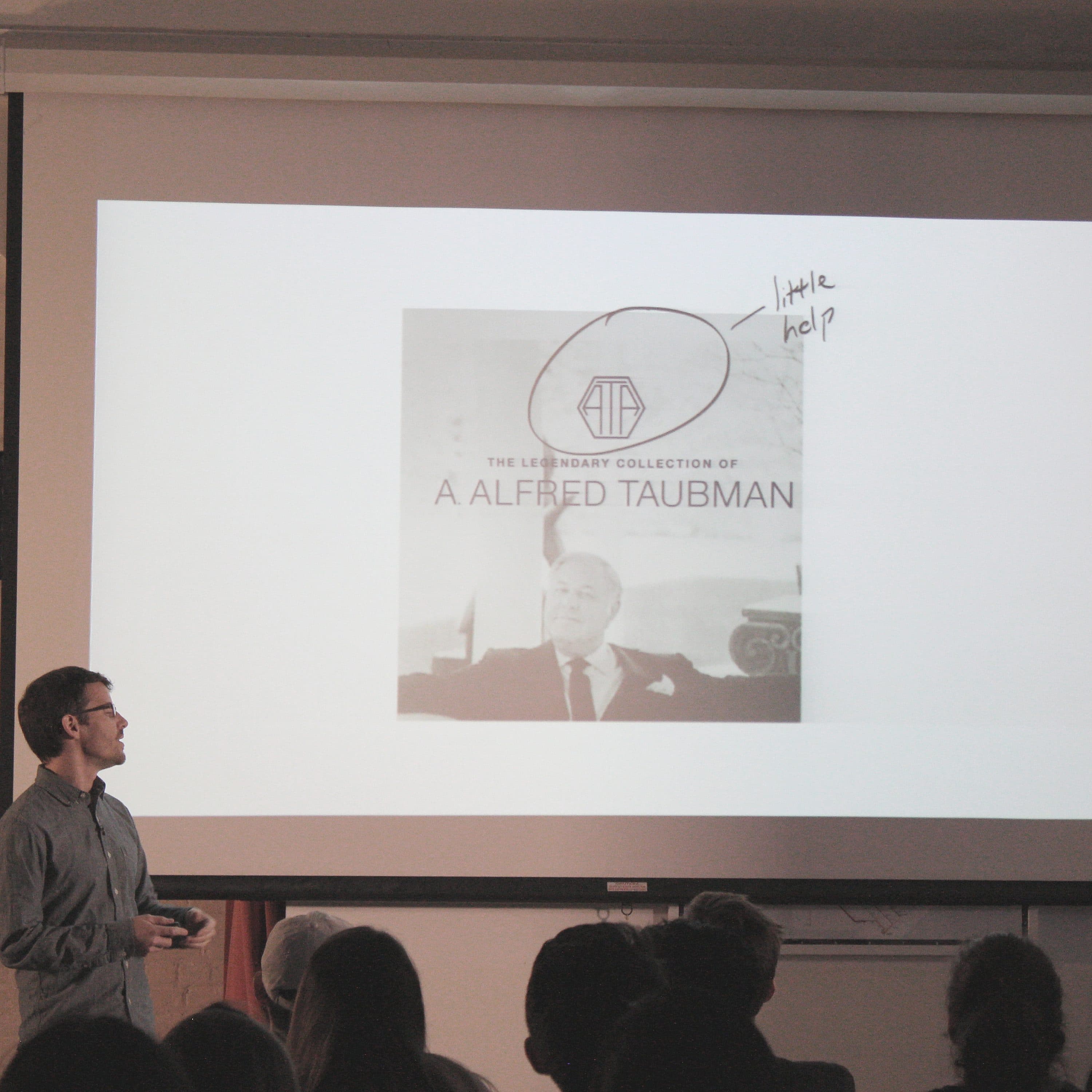 A man is standing in front of a projection screen, giving a presentation. The screen displays an image labeled "The Legendary Collection of A. Alfred Taubman." There are framed pictures on the wall behind him, and an audience is seated, watching the presentation.