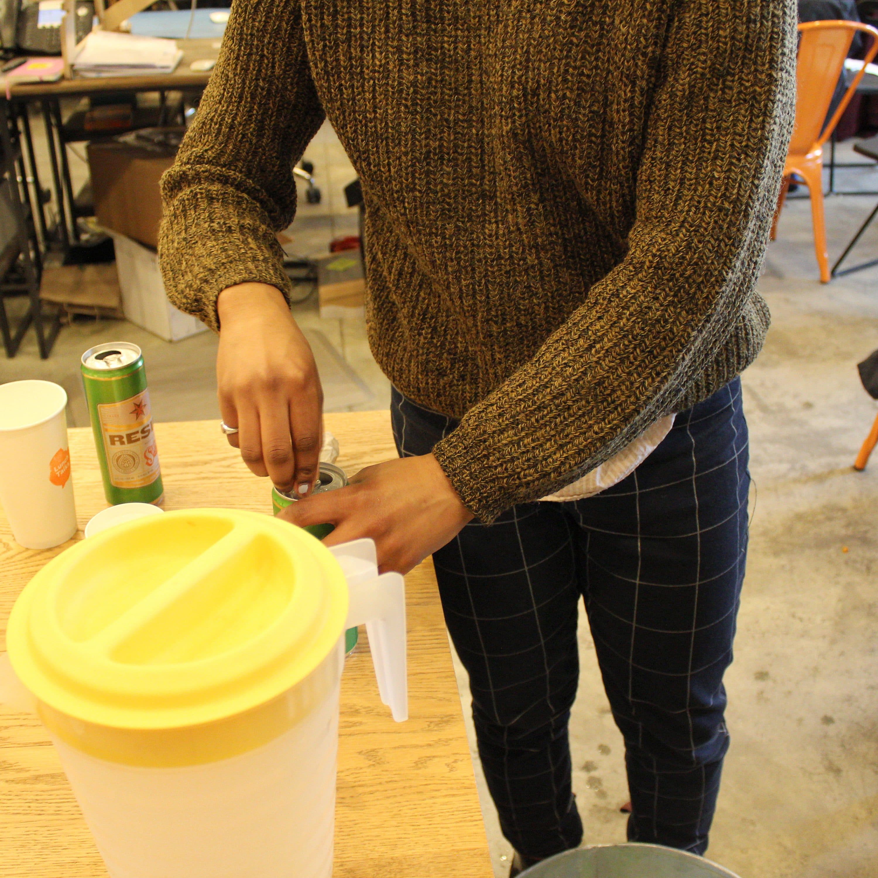 A person in a brown sweater is pouring a beverage from a yellow-lidded container into a transparent cup. There's a green can and several empty cups on the wooden table. The scene appears to be in a casual indoor setting with chairs and other items in the background.
