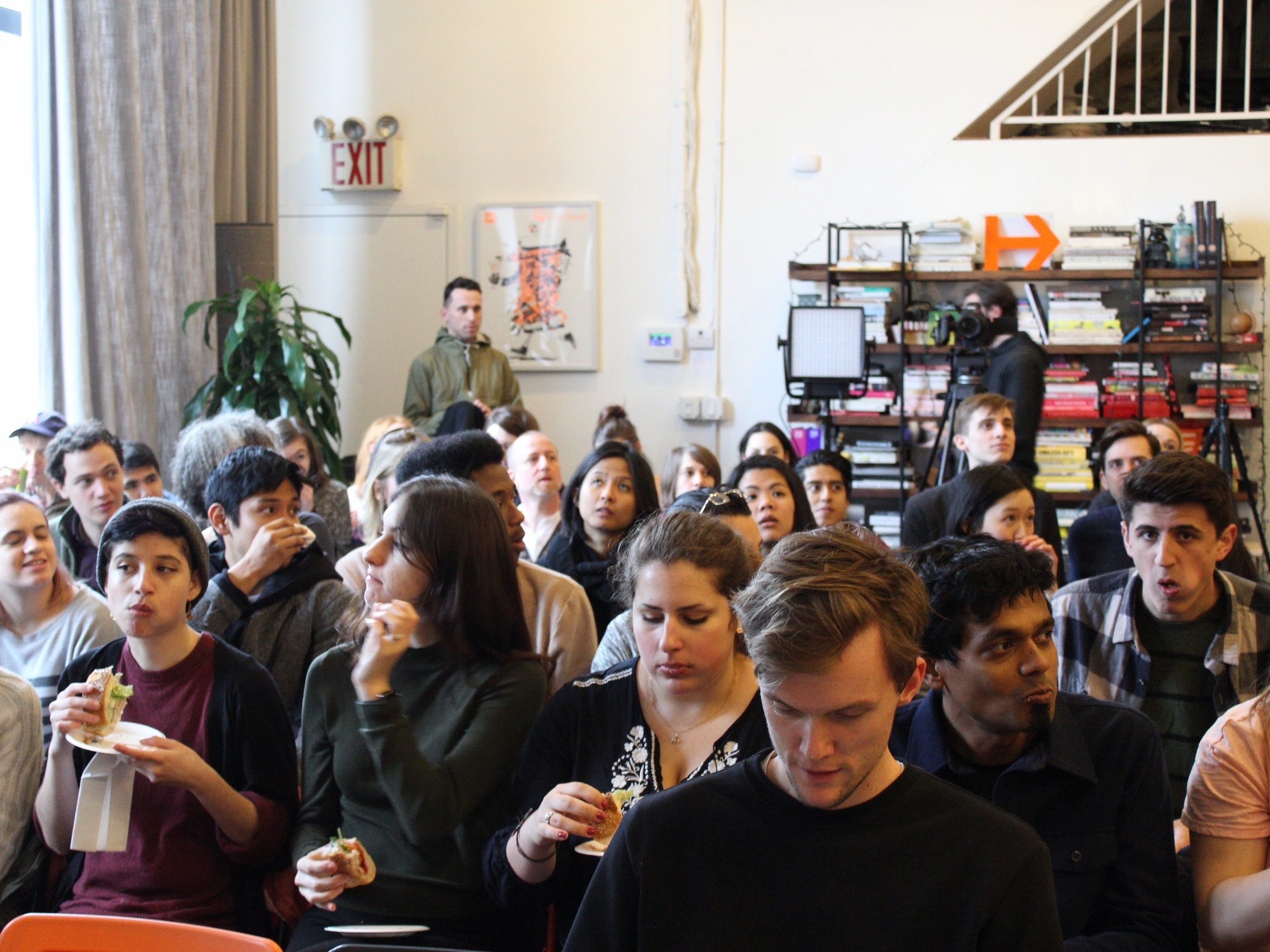 A diverse group of people sit closely together in a room, attentively listening to something off-camera. Some hold food and drinks, possibly indicating a casual or social event. In the background, there are shelves of books and a staircase.
