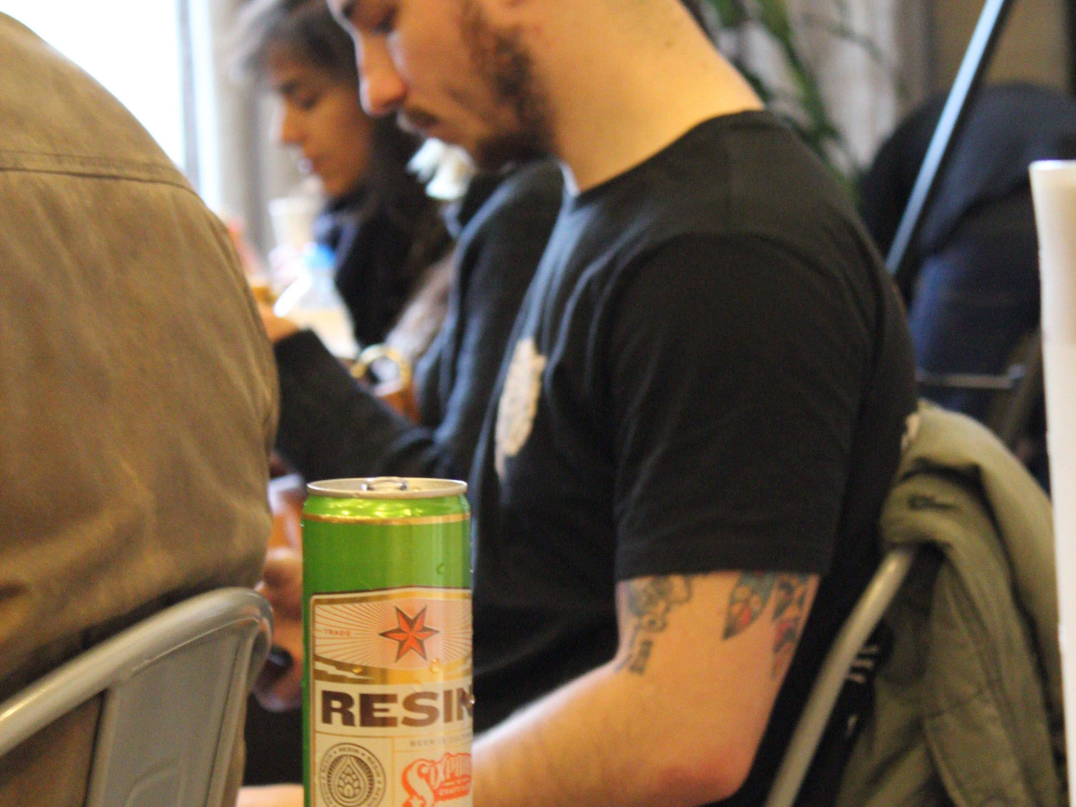 A person wearing a gray beanie and black t-shirt sits in a room with another individual. In the foreground, there is a green can of beer labeled "Resin" on a wooden table. A jacket is draped over the back of the person's chair. A plant is visible in the background.