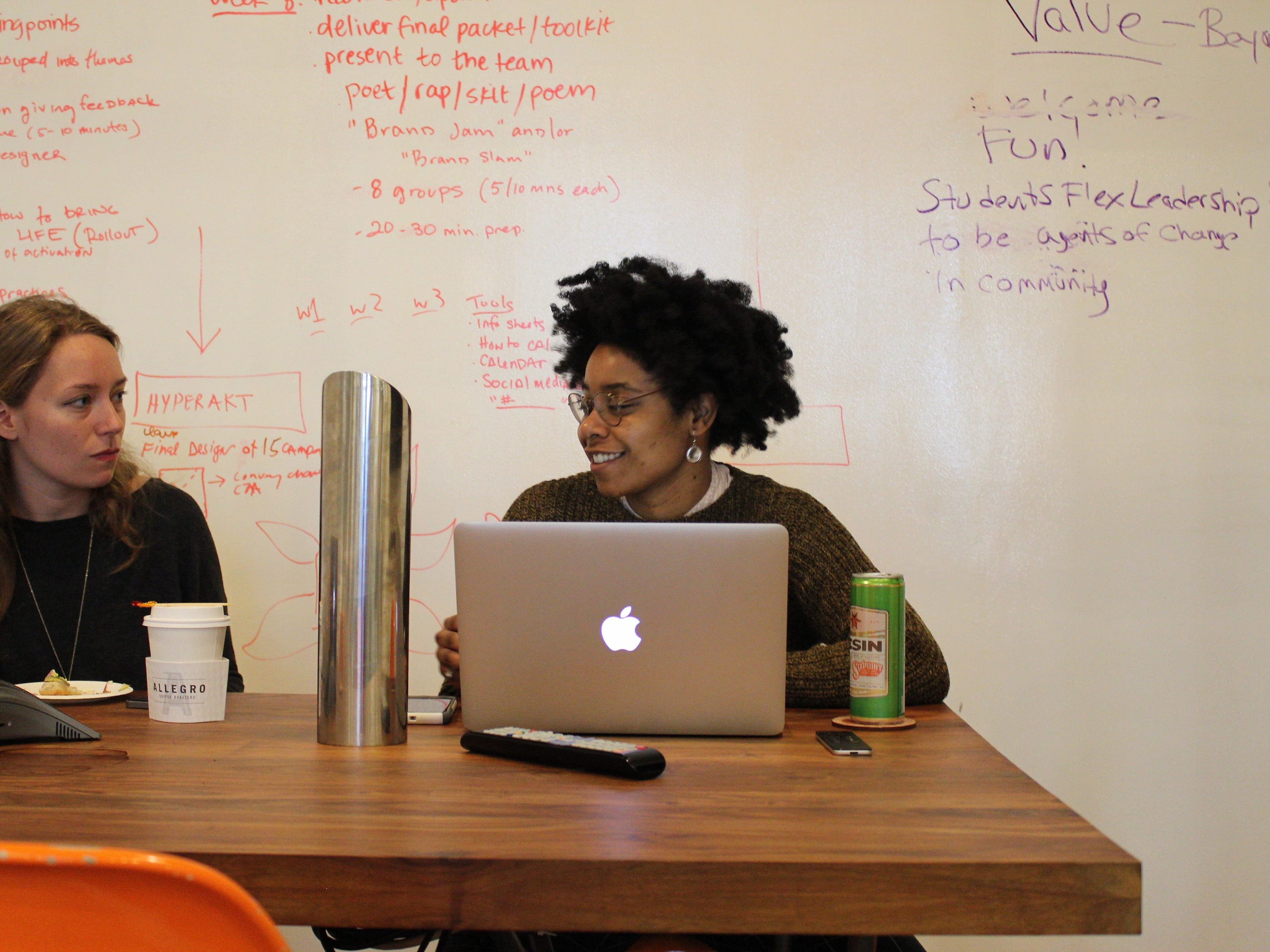Two people sit at a wooden table in front of a whiteboard covered in red and black text. One person with curly hair and glasses uses a MacBook, smiling. The other looks at her, holding a plate. The table has a metal water bottle, coffee cups, and a drink can.
