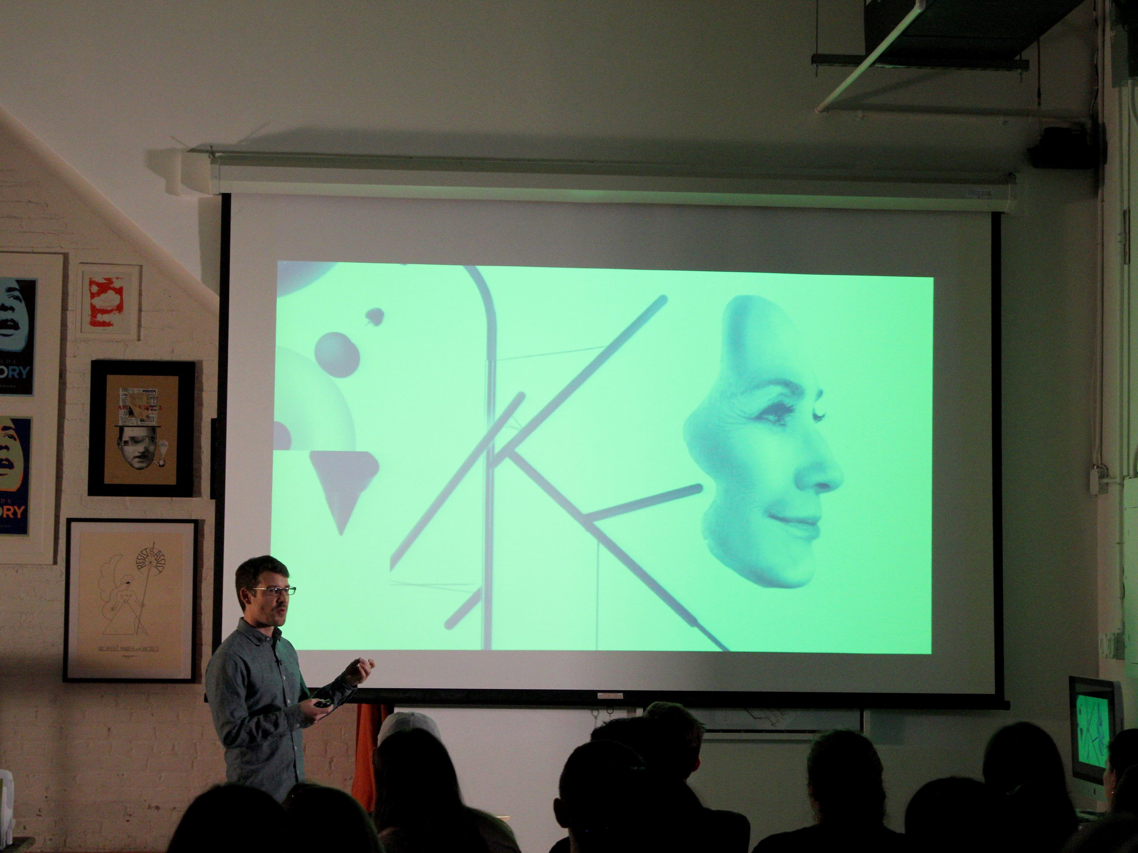 A person is standing and presenting in front of a screen displaying an abstract image with overlapping faces and geometric shapes in green hues. The room has several framed posters on the walls, and an audience is seated facing the presenter.