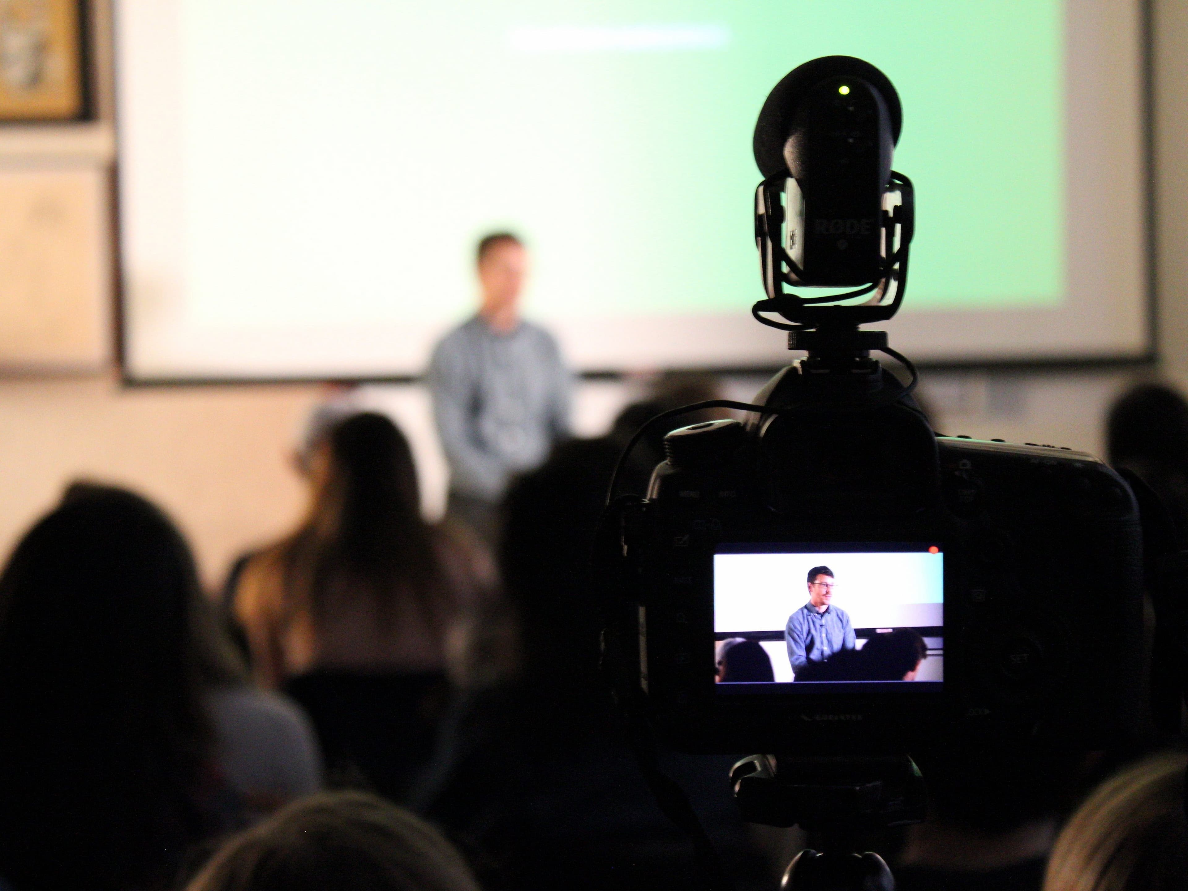 A speaker is presenting in front of an audience, seen out of focus in the background. A camera in the foreground is recording the event, with the speaker clearly visible on the camera’s display screen. The room appears to be dimly lit.