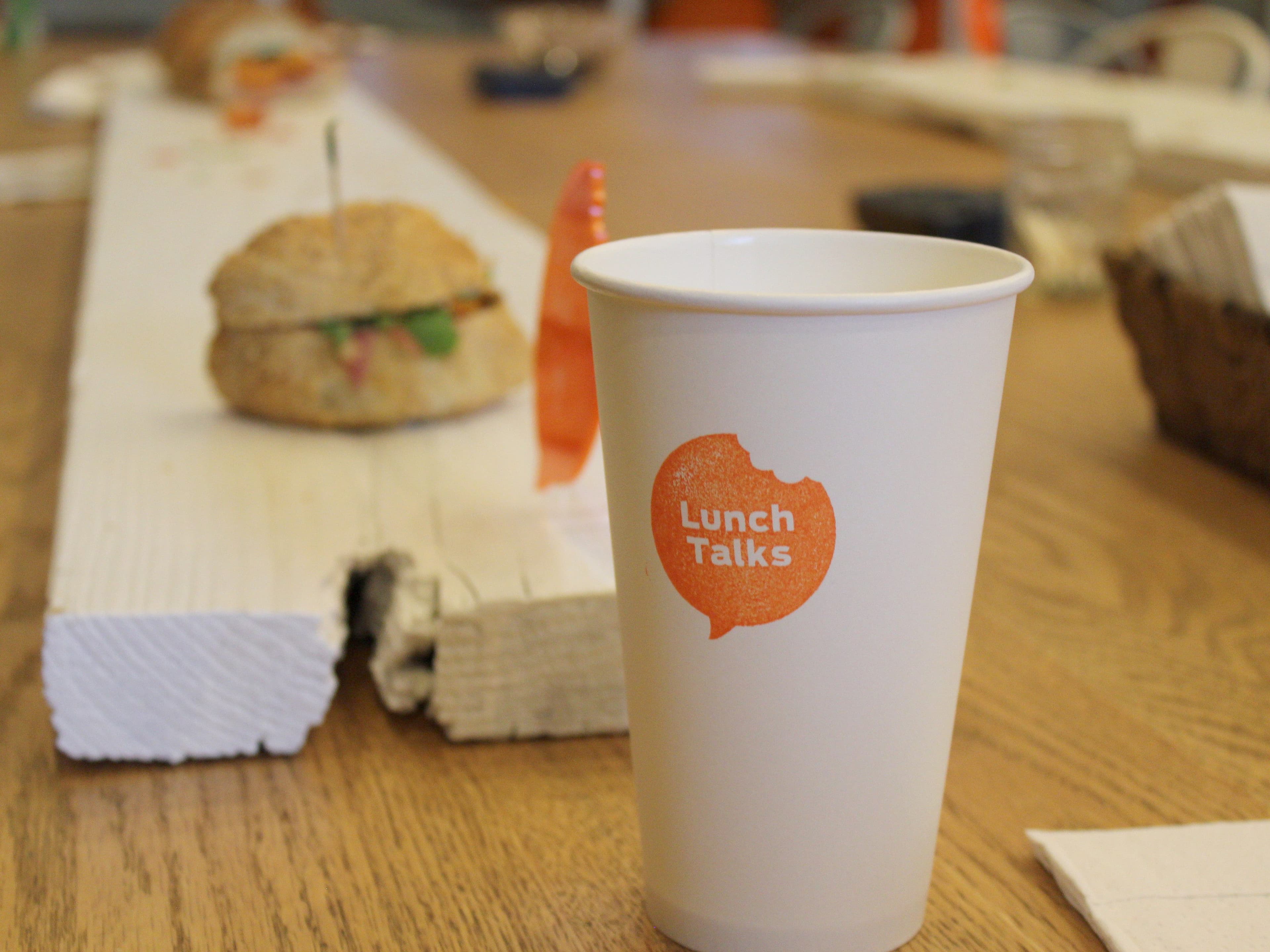 A white paper cup with an orange logo reading "Lunch Talks" sits on a wooden table. In the background, there is a sandwich on a white plank and a small plastic orange dinosaur toy, slightly out of focus. The setting suggests an informal gathering or discussion over lunch.