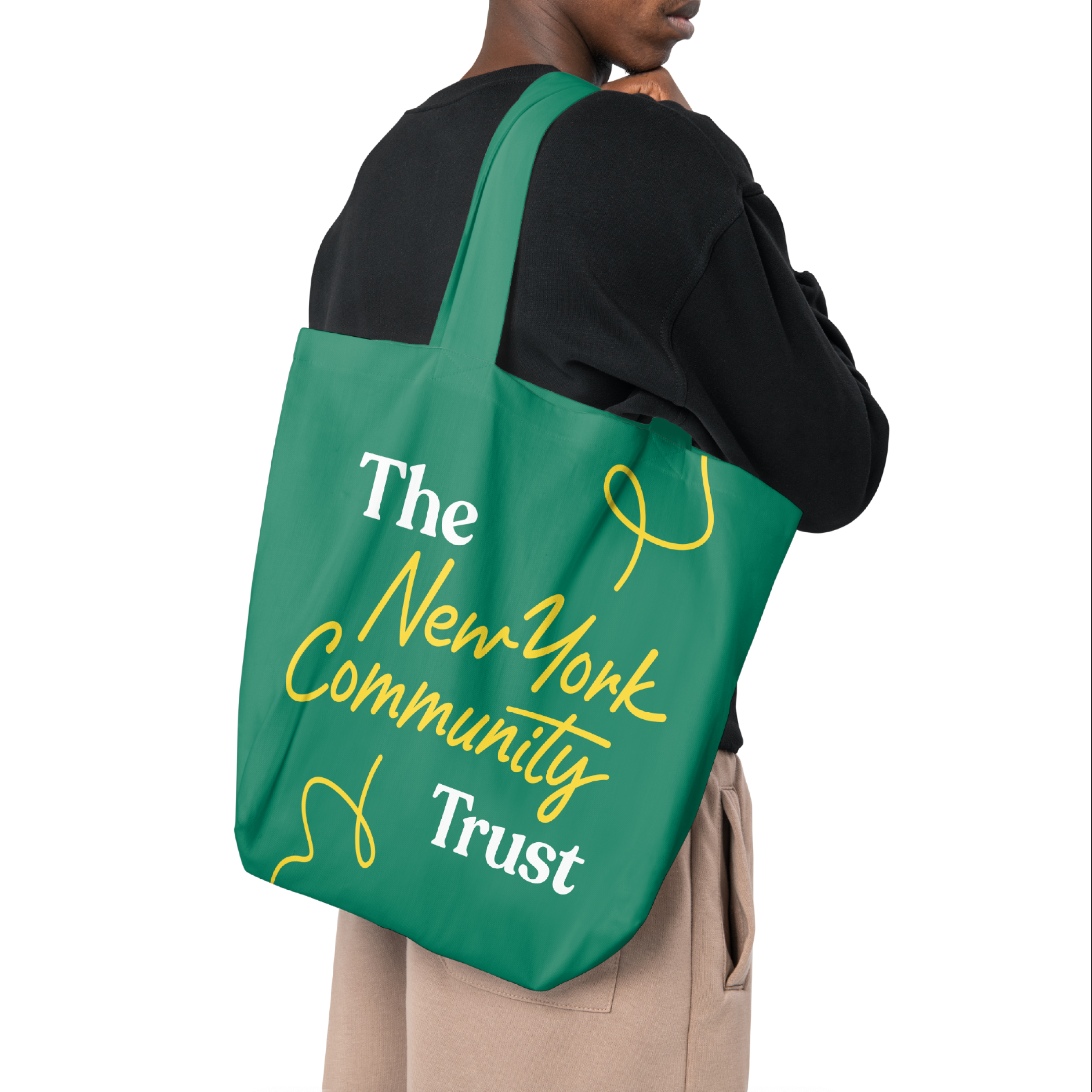 A person in a black shirt and beige pants carries a green tote bag with "The New York Community Trust" written in white and yellow text. The tote bag features a simple, stylish design with yellow cursive lines.