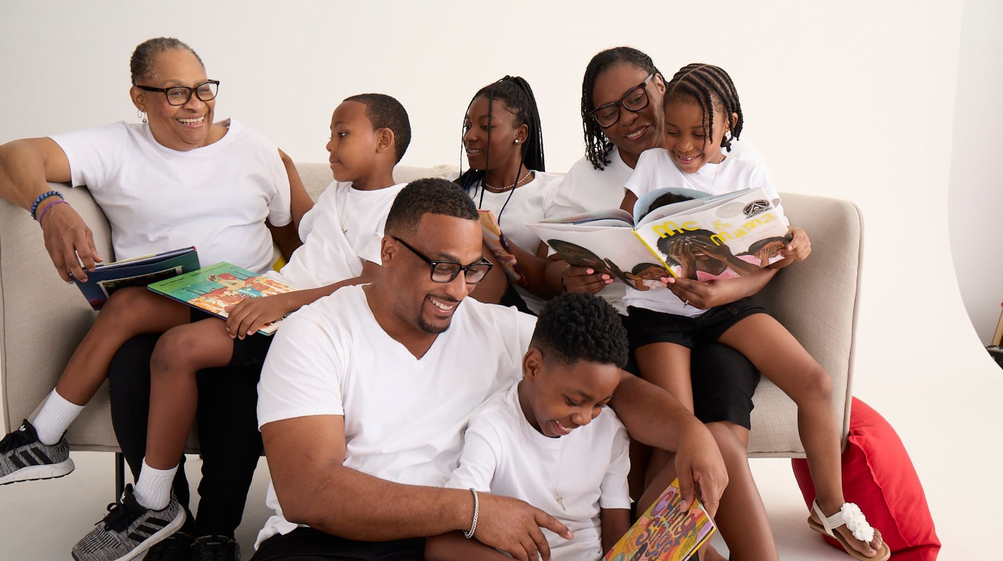 A joyful family sits together on a sofa, dressed in matching white shirts. The adults and children read colorful books while smiling and engaging with one another. The scene reflects a warm, light-hearted moment of shared activity and togetherness.