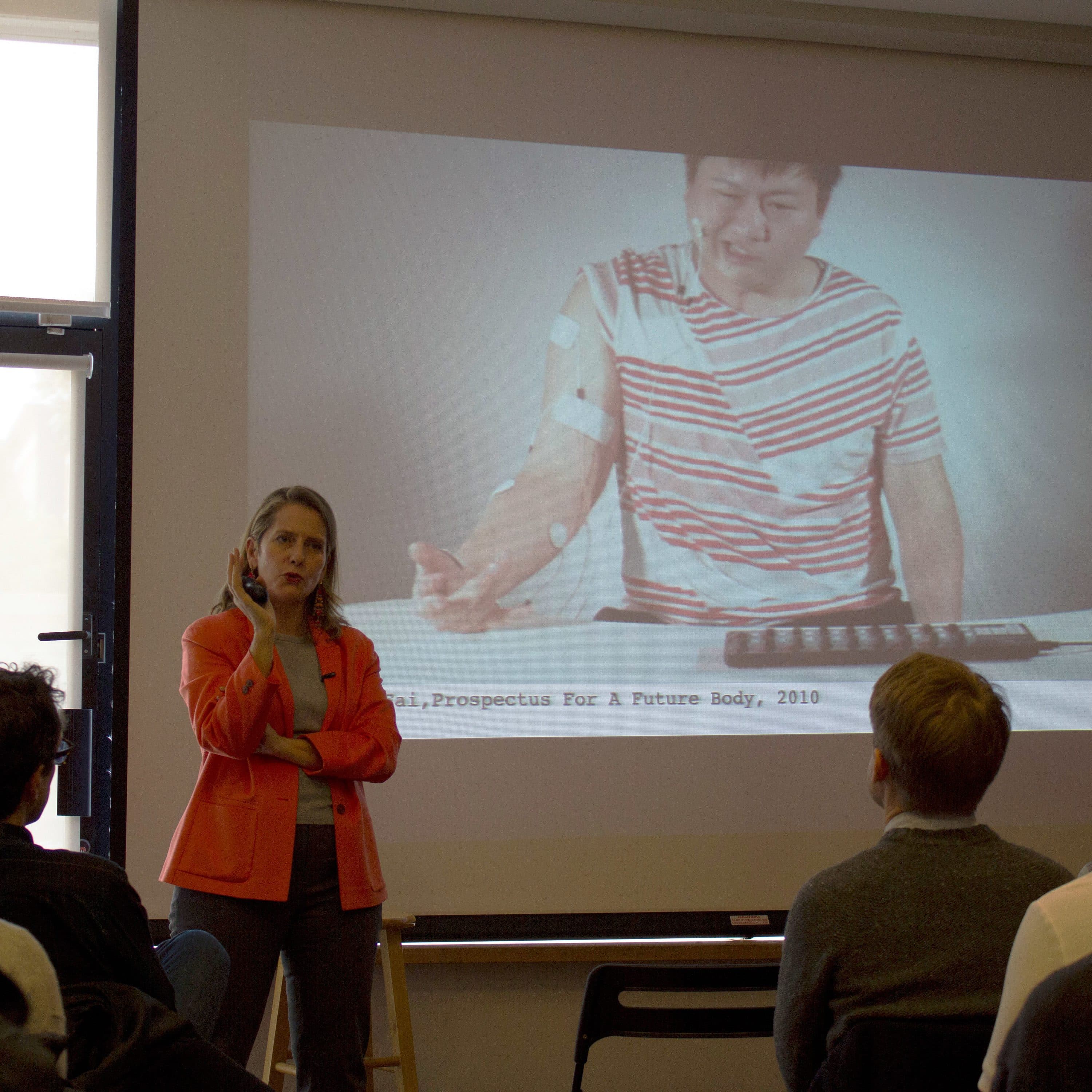 A woman standing in front of a projected screen is speaking to an audience. The screen displays an image of a person with electrodes on their arm, interacting with a keyboard. The indoor setting has a mix of natural and artificial lighting, with an open door on the left.