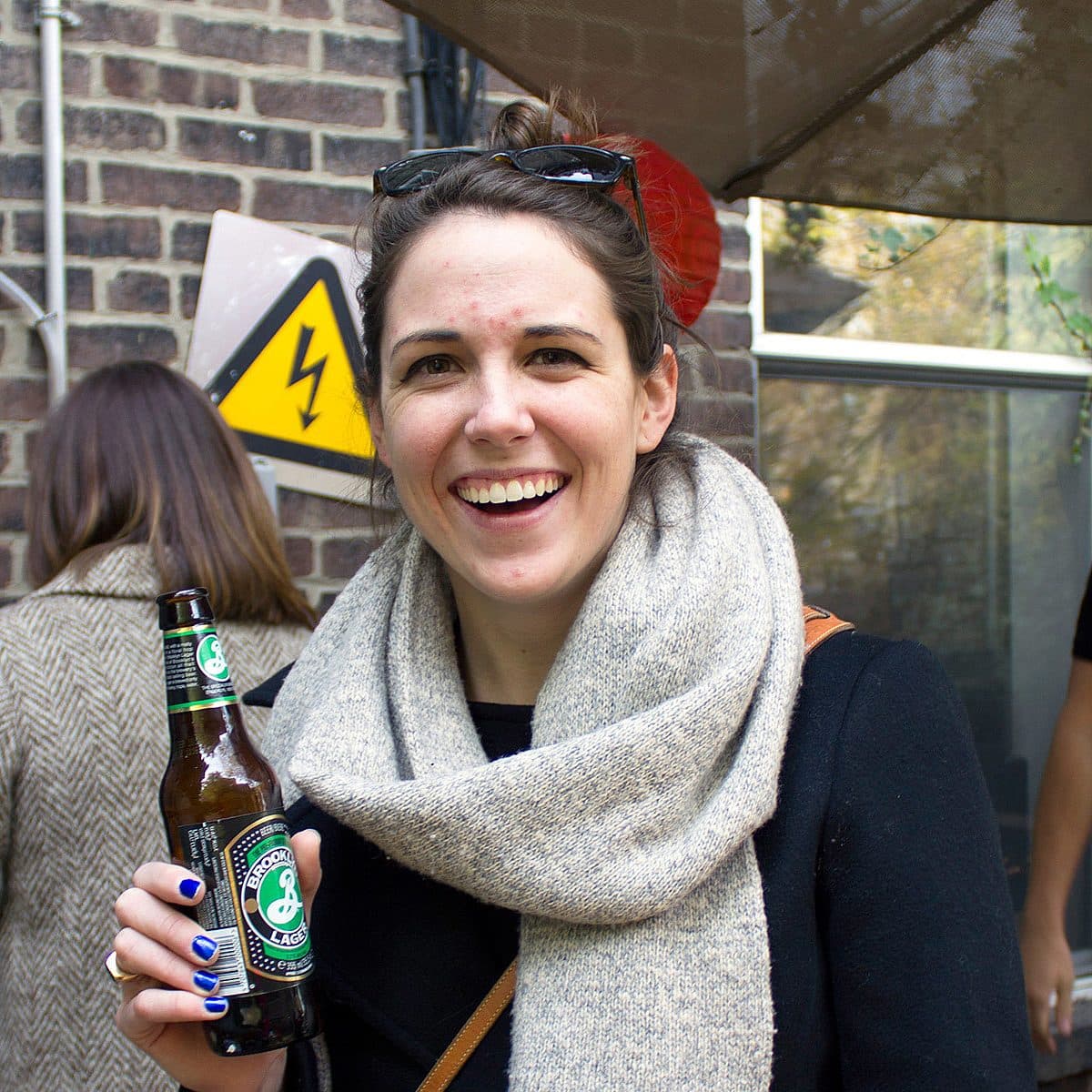 A woman smiles while holding a bottle of beer. She's wearing a dark jacket and a beige scarf. In the background, there are other people and a sign with a lightning bolt. The setting appears to be outdoors.