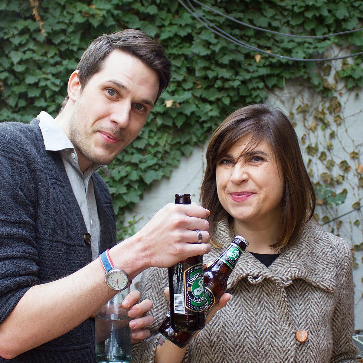A man and a woman are standing outside in front of a wall covered in ivy. Both are holding bottles of lager, with the man holding an additional glass. The man is wearing a black sweater and a watch; the woman is wearing a brown coat. They are smiling at the camera.
