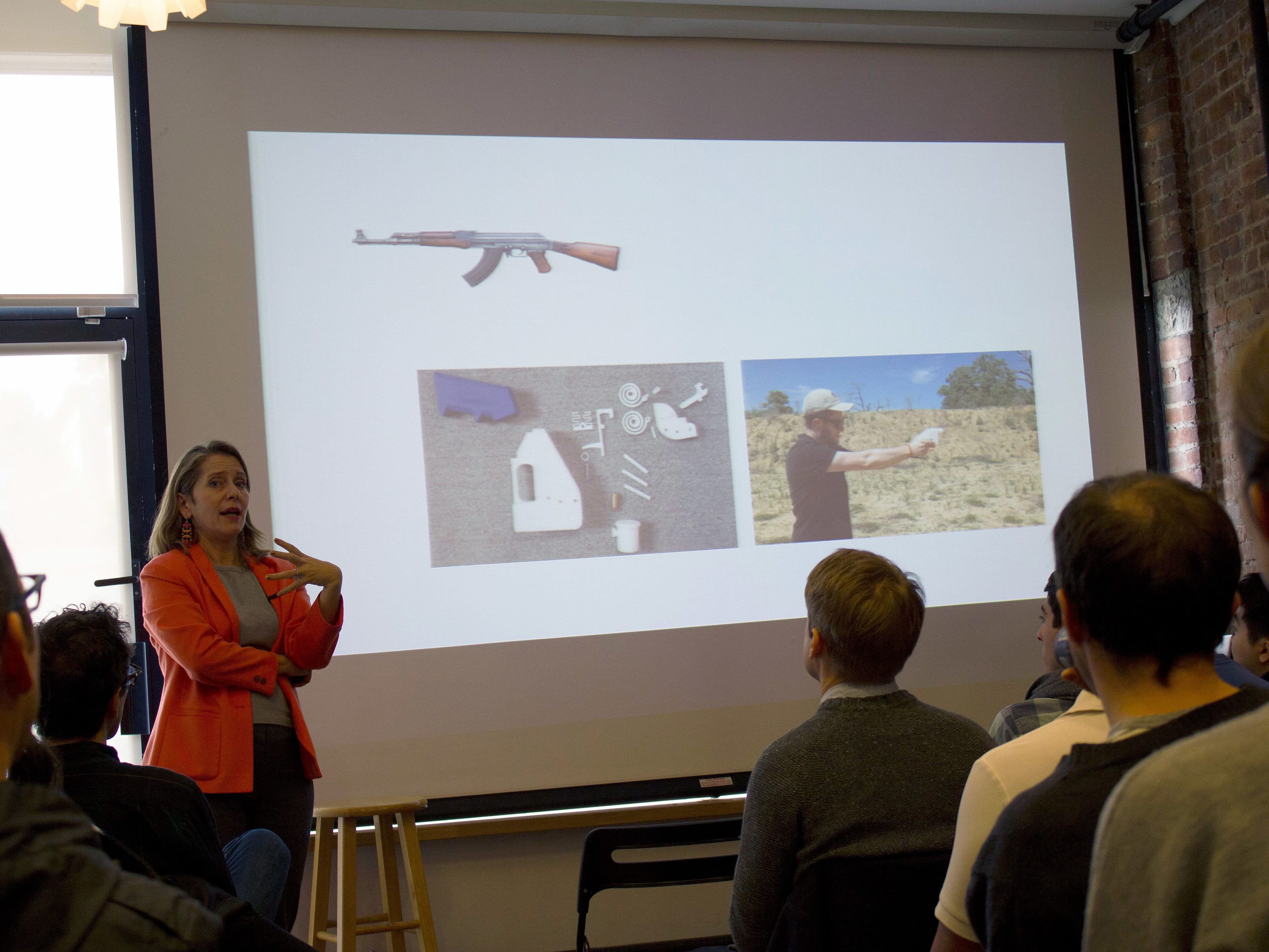 A woman in an orange blazer is speaking in front of a group of people in a classroom setting. On the screen behind her are images of a rifle, 3D-printed gun parts, and a person shooting a firearm outdoors. The audience is seated and attentively listening.