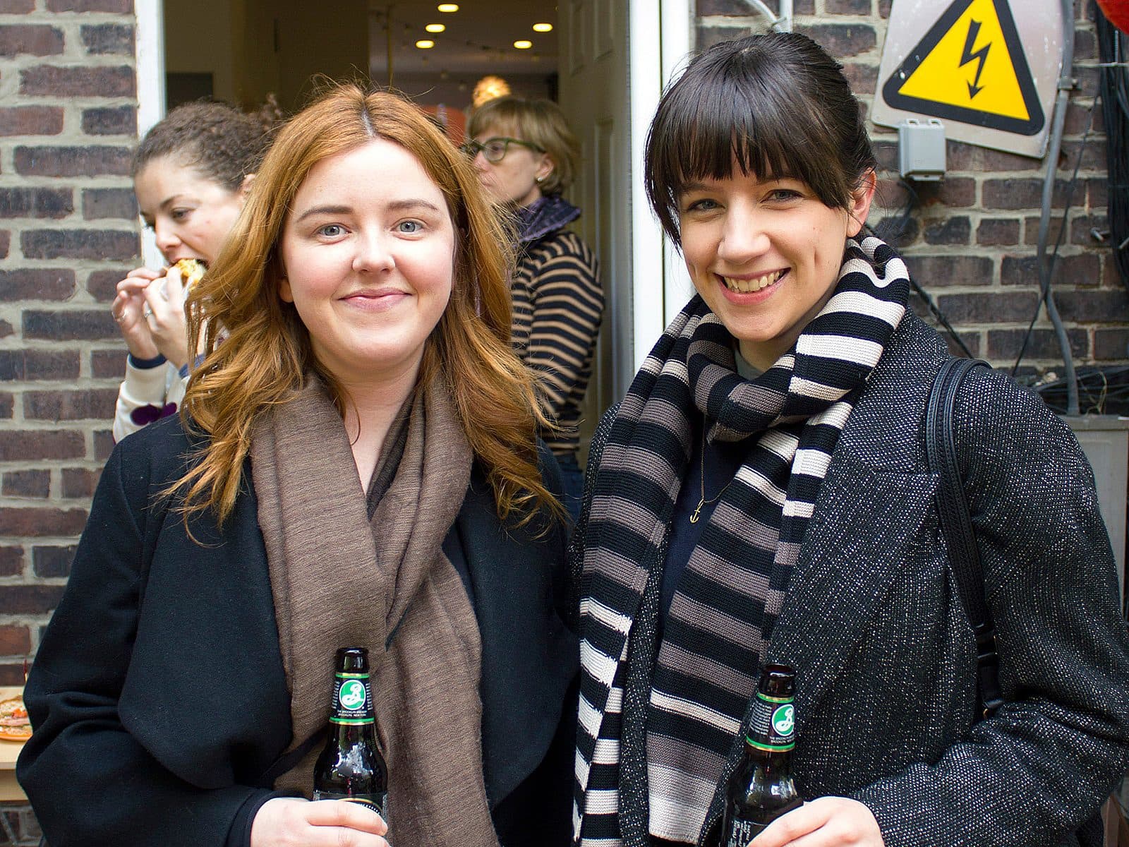 Two women, both holding bottles of beer, pose for a photo outdoors against a brick wall with caution stickers. One woman has long red hair, and the other has dark hair in a ponytail with bangs, both smiling. In the background, two people are partially visible, one eating pizza.