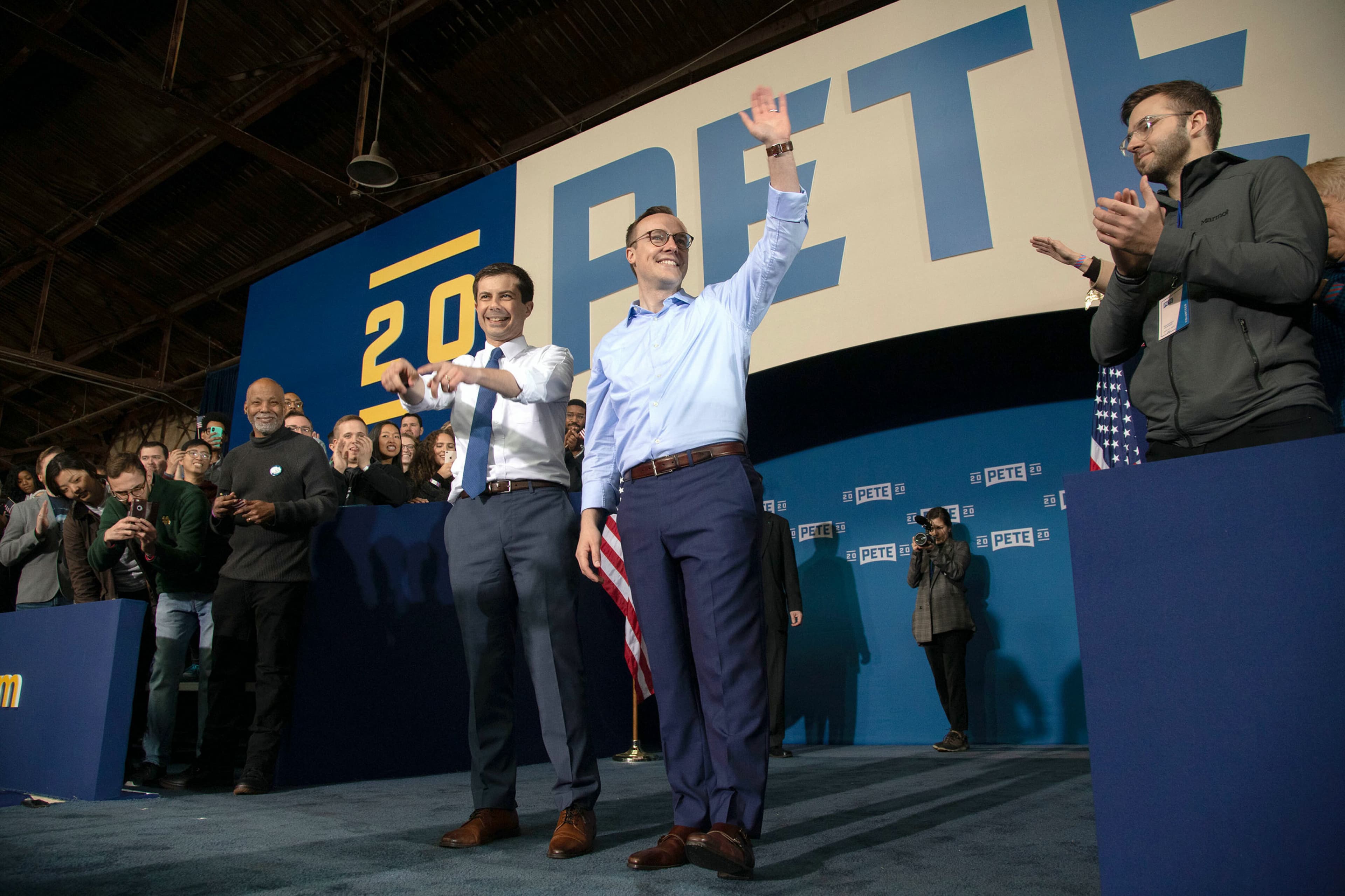 Two people standing on a stage wave to a crowd. The person on the right holds an American flag. Behind them is a large sign with the name "PETE" and the year "2020." Several people on the stage and in the audience clap and smile.