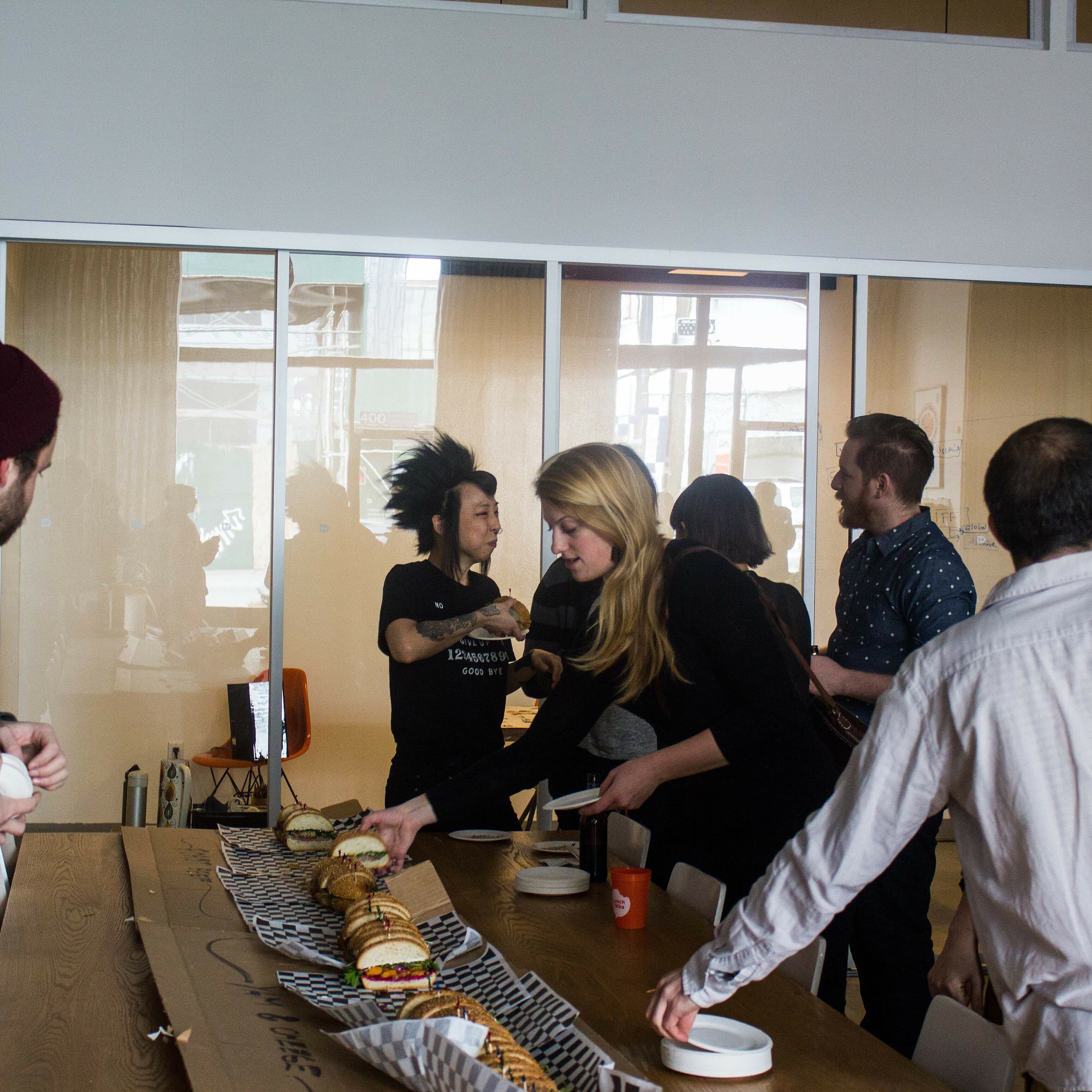 A group of people in a modern office are gathered around a table with assorted sandwiches. Two individuals are reaching for food, while others engage in conversation. One person wears a red beanie, and another has a distinctive hairstyle. Glass partitions are in the background.