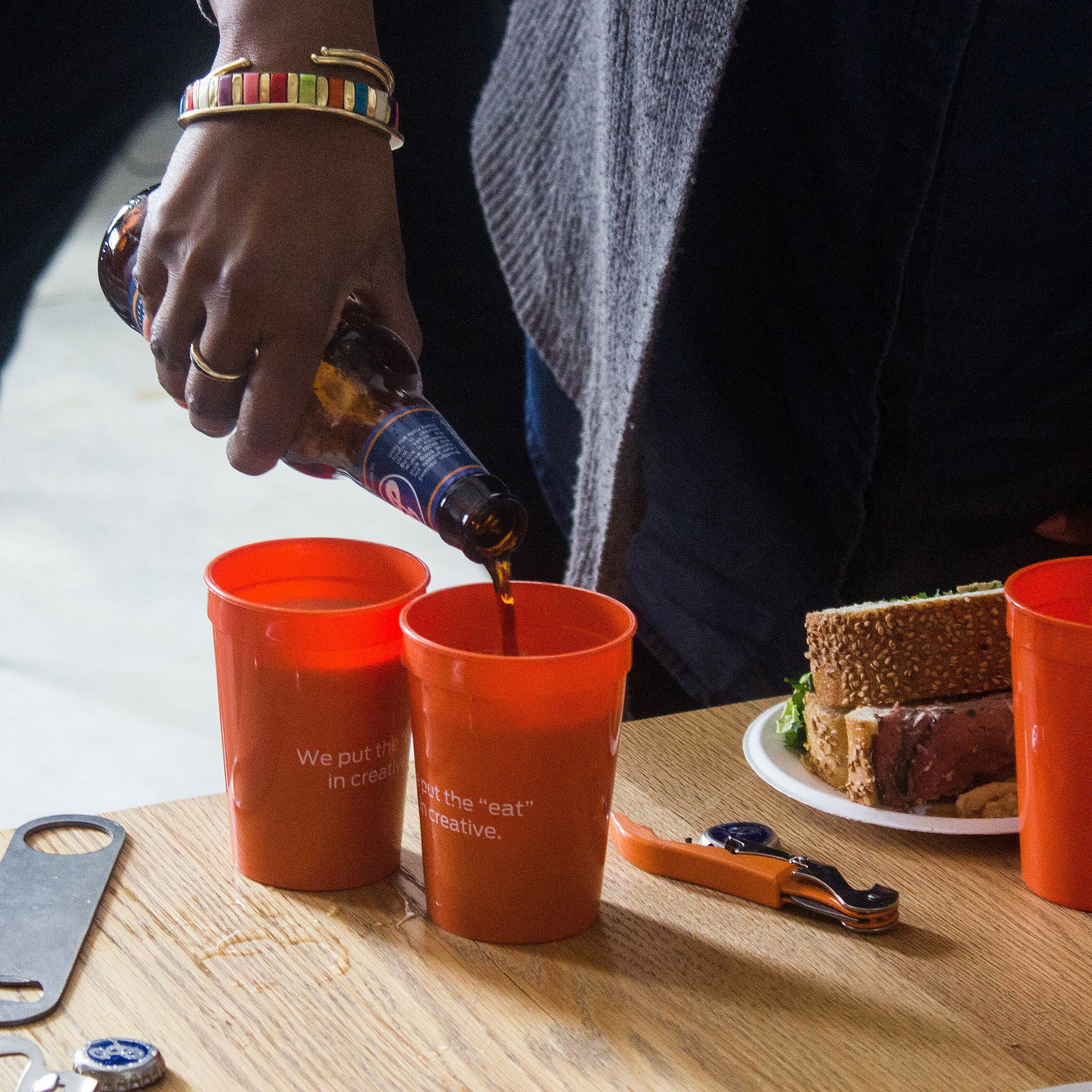 A person is pouring a dark beverage from a bottle into one of three orange cups on a wooden table. The table also has a plate with food, a bottle opener, and metal utensils. The cups have white text and the Lunch Talks logo.