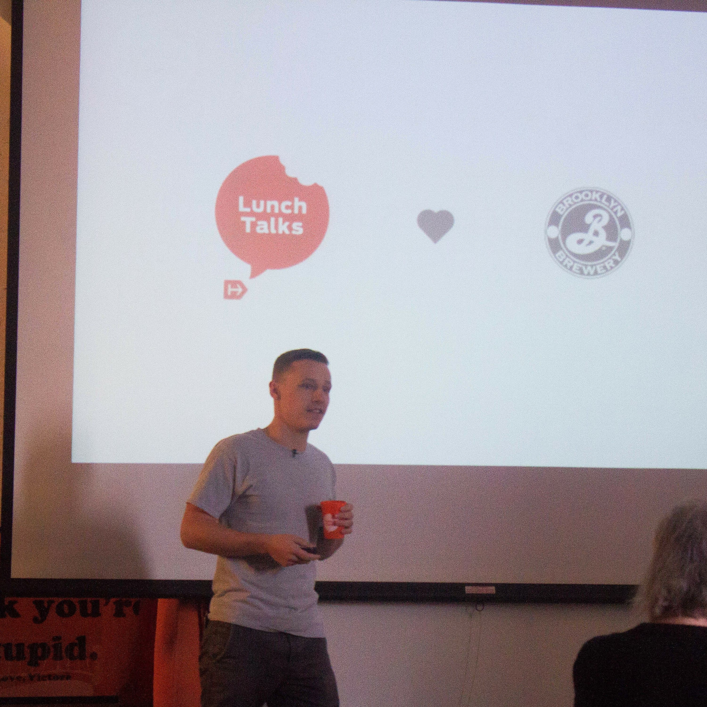 A man in a grey t-shirt stands in front of a projection screen holding a microphone and a red cup. The screen shows logos for "Lunch Talks," "Brooklyn Brewery," and "D Co. Design" with a heart symbol between them. People are seated facing him.
