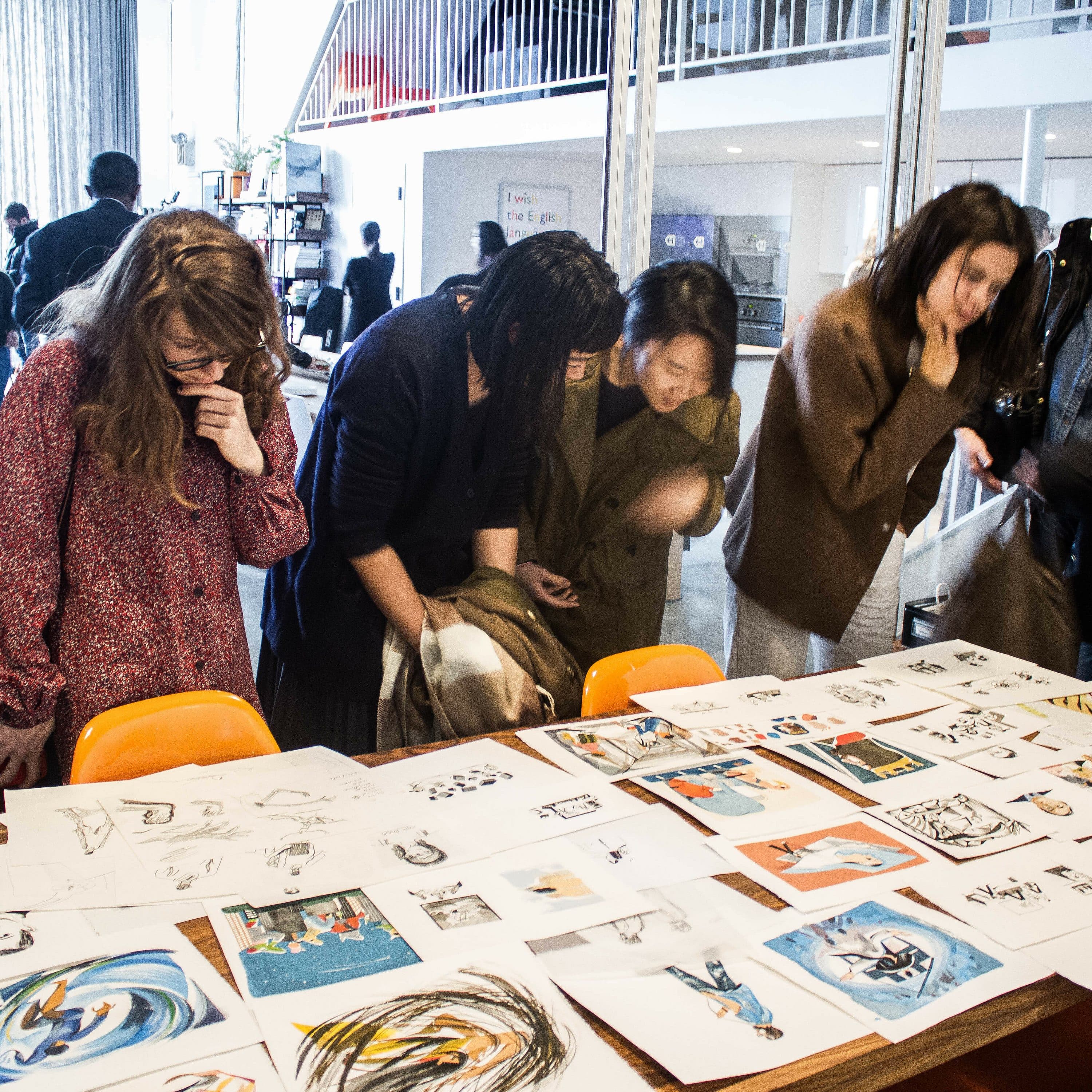 A group of people is gathered around a table filled with various sketches and artwork. They seem to be closely observing and discussing the pieces. The setting appears to be a well-lit, creative studio space with other individuals in the background.