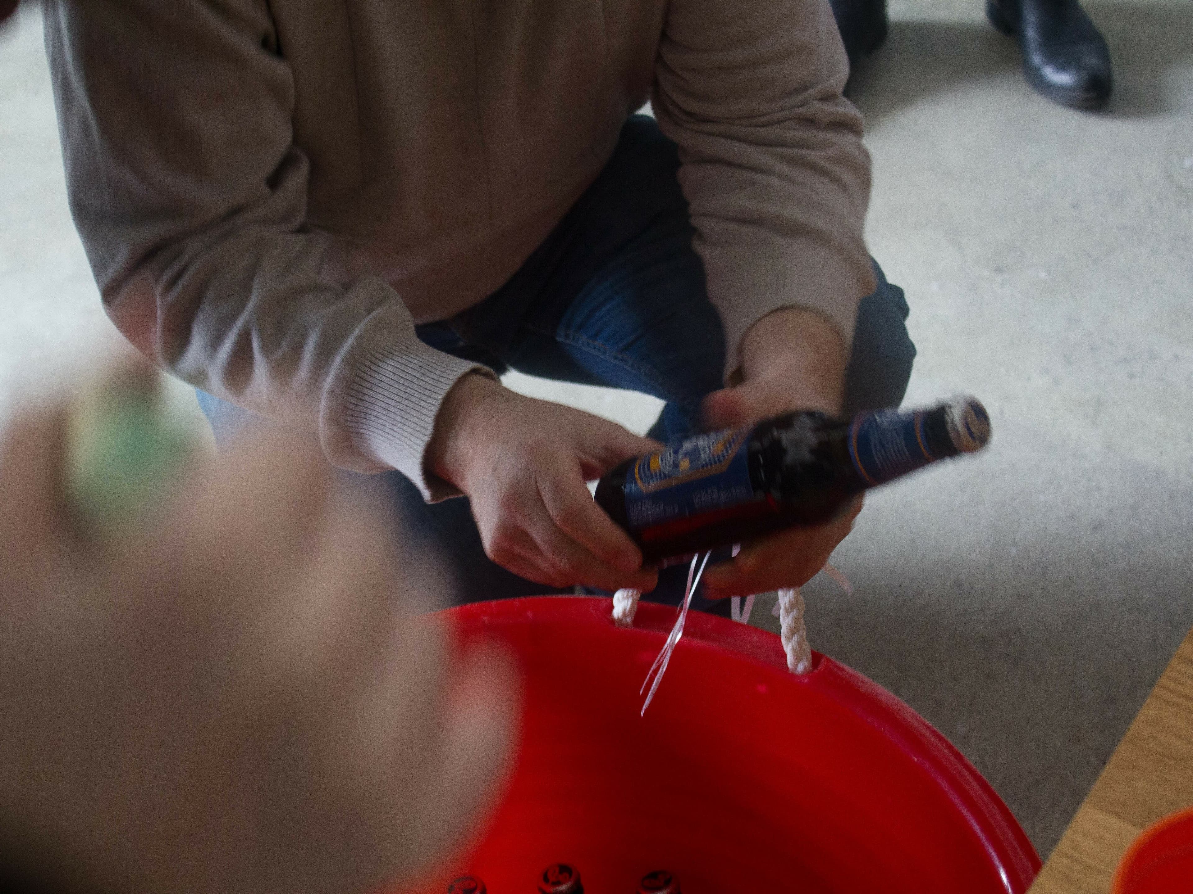 A person in a light-colored sweater and jeans crouches beside a large red bucket, holding a dark-colored bottle with a label. Another person is partially visible in the foreground, reaching towards the bucket. The scene appears to be indoors.