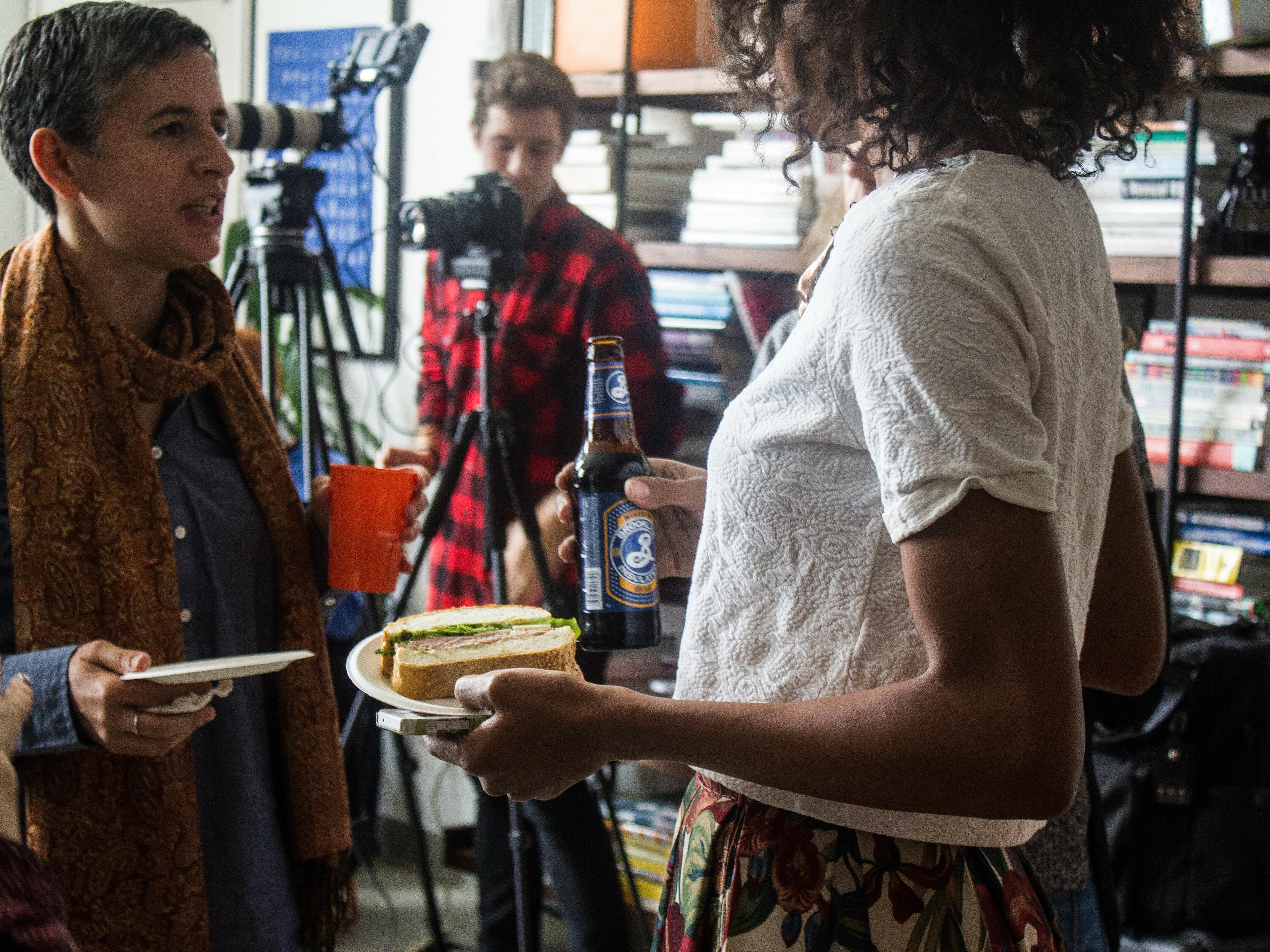People are gathered in an indoor setting with shelves filled with books and objects in the background. A woman is holding a plate with a sandwich and a bottle of beer, while another person holds a red mug. A man in a red flannel shirt operates a camera in the background.