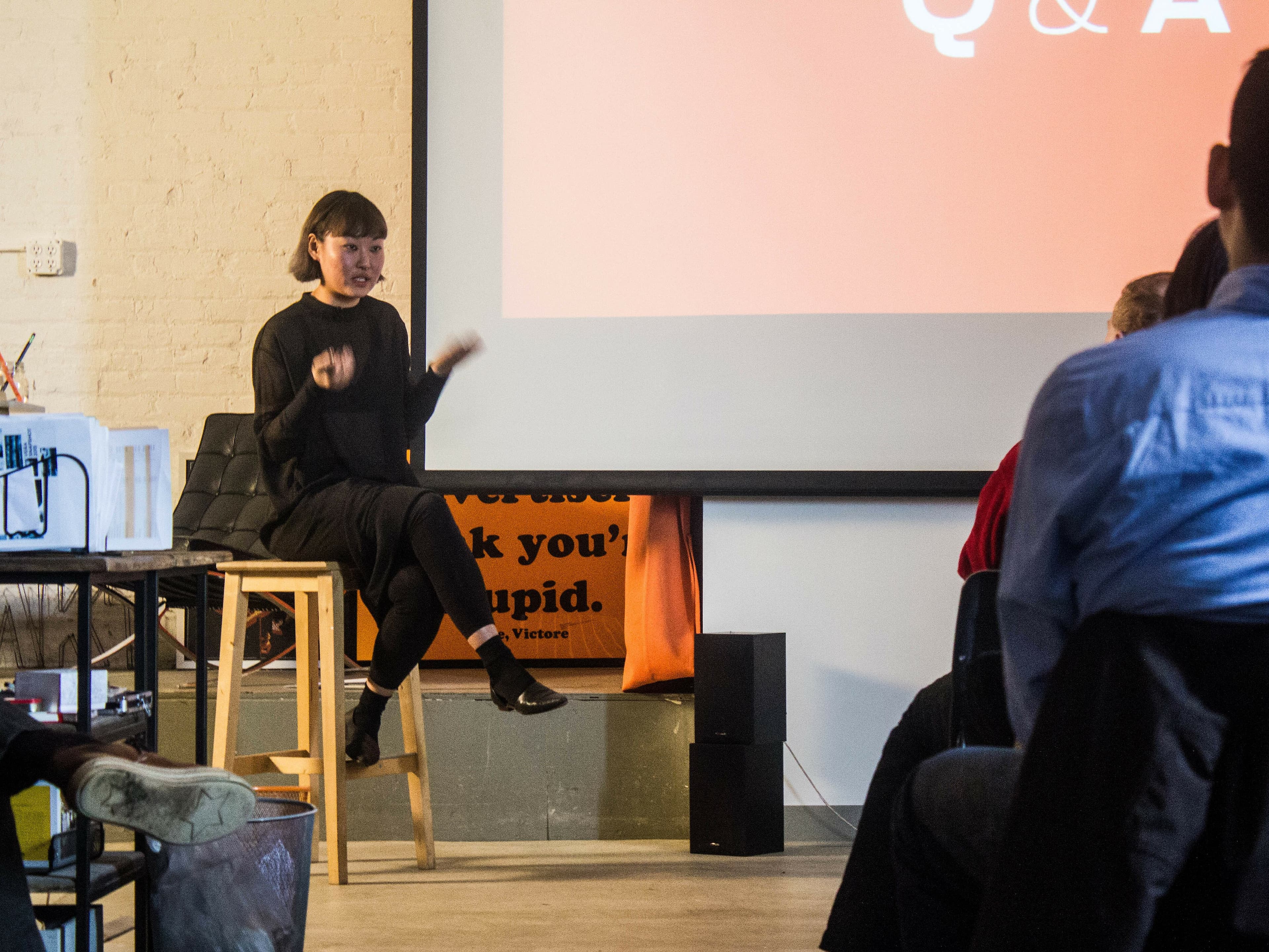 A person wearing black sits on a stool in front of a group, gesturing with their hands. Behind them, a large screen displays "Q & A" on an orange background. The setting appears to be an indoor event or lecture. Audience members are seated facing the speaker.