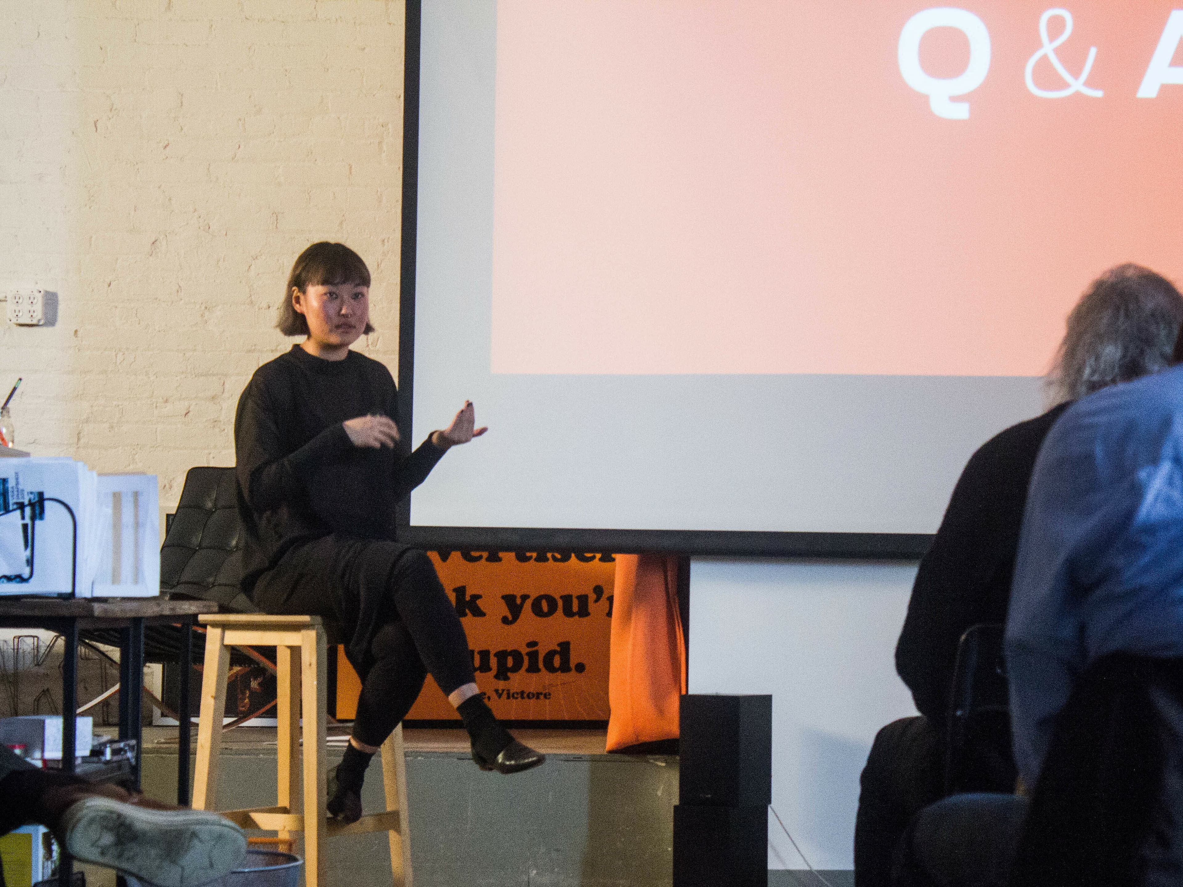 A person sits on a stool giving a presentation in front of a large screen displaying "Q & A." The setup suggests a casual, creative environment with a 3D printer visible on the left side. The audience is attentively listening.