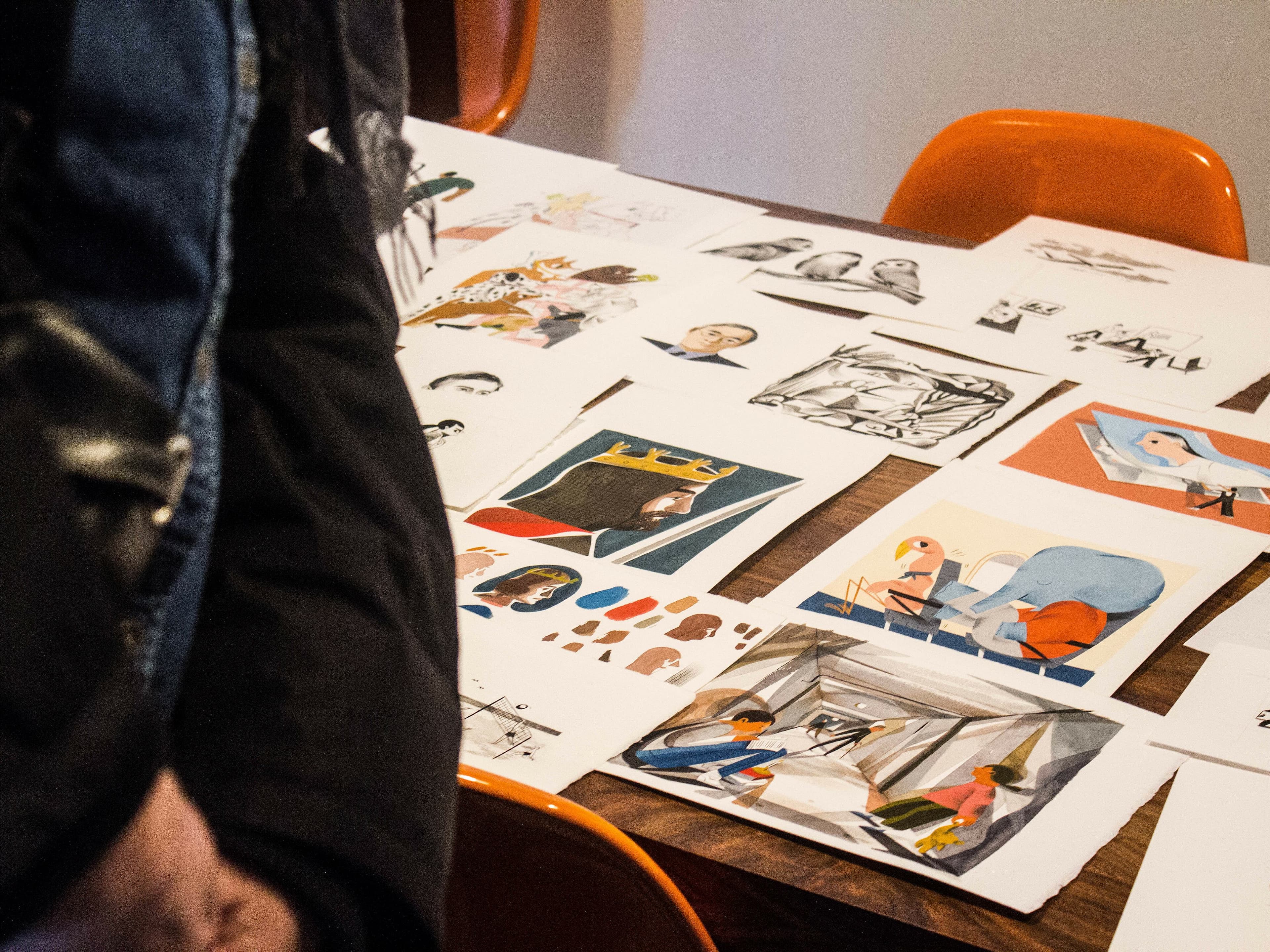 A table covered with various colorful drawings and illustrations, some depicting people and scenes, is being observed by a person in a black jacket. Bright orange chairs surround the table. The focus is on the artwork spread across the table.