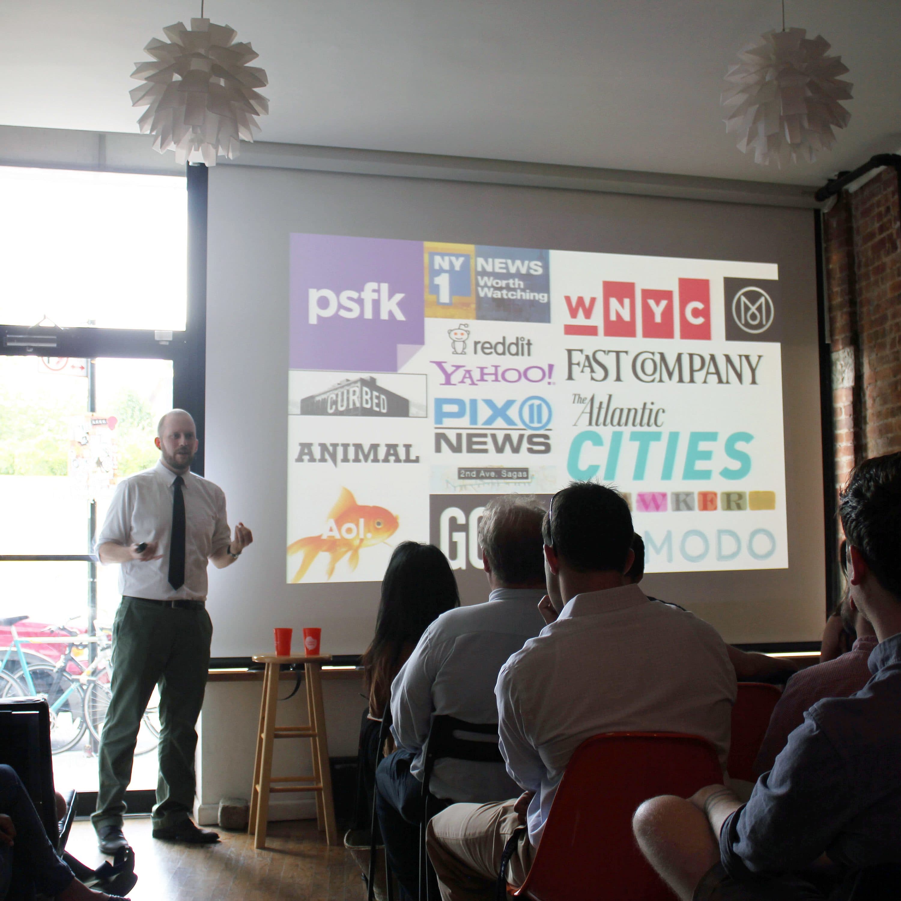 A man stands at the front of a room giving a presentation to a seated audience. A projector screen behind him displays various media logos such as Fast Company, Atlantic Cities, and Yahoo!. The room has a modern design with brick walls and flower-shaped hanging lights.
