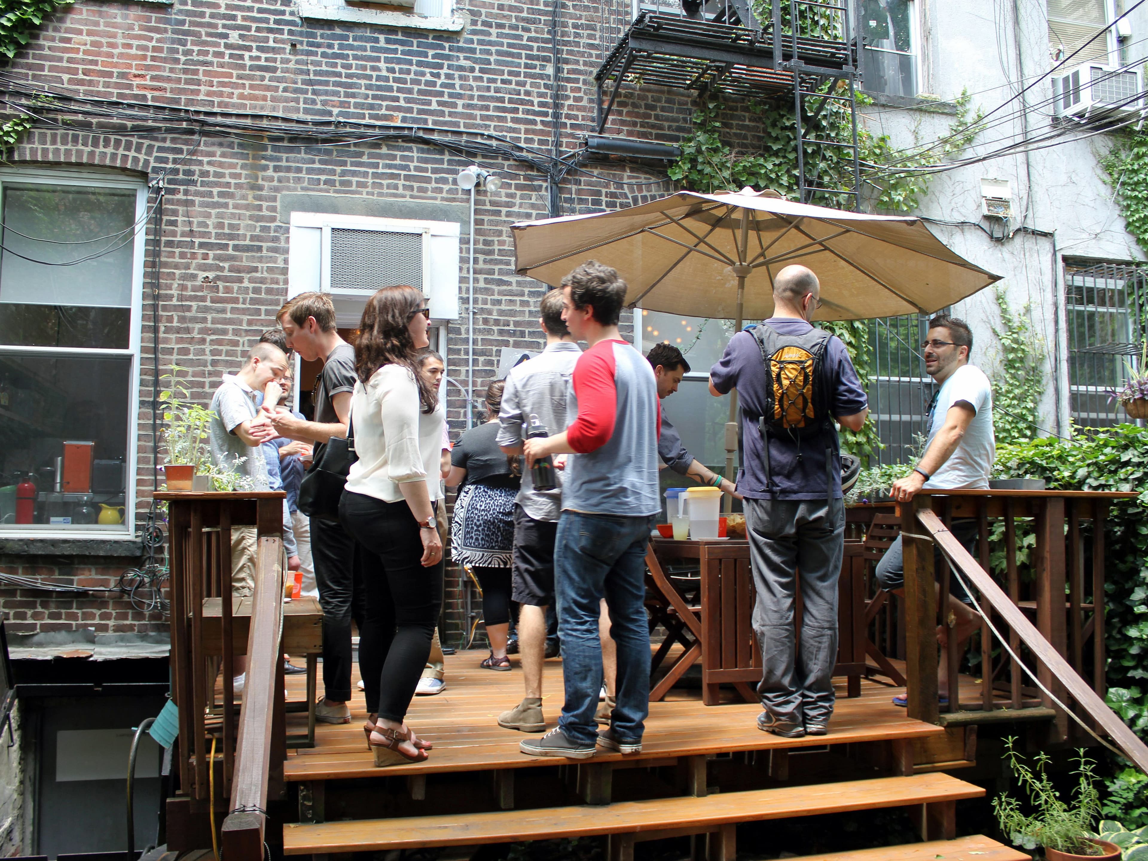 A group of people are gathered on a wooden deck in a backyard. They are socializing under a large umbrella on a sunny day. The surrounding buildings have brick and ivy-covered walls. Some people are holding drinks, and there is a table with refreshments.
