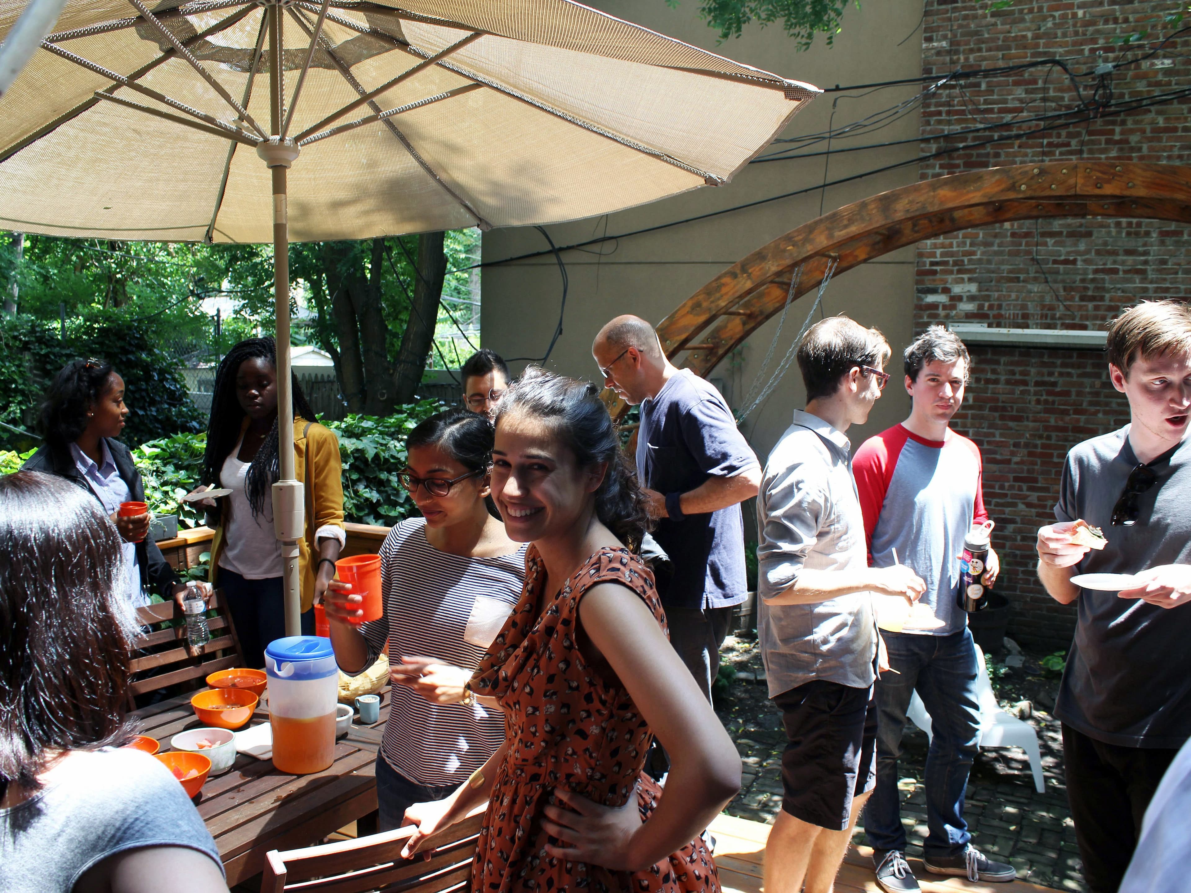 A group of people gather outdoors under a large umbrella in a garden setting. They are engaged in conversation, holding drinks, and eating food from a table filled with snacks. Trees and a brick wall with a wooden archway are visible in the background.