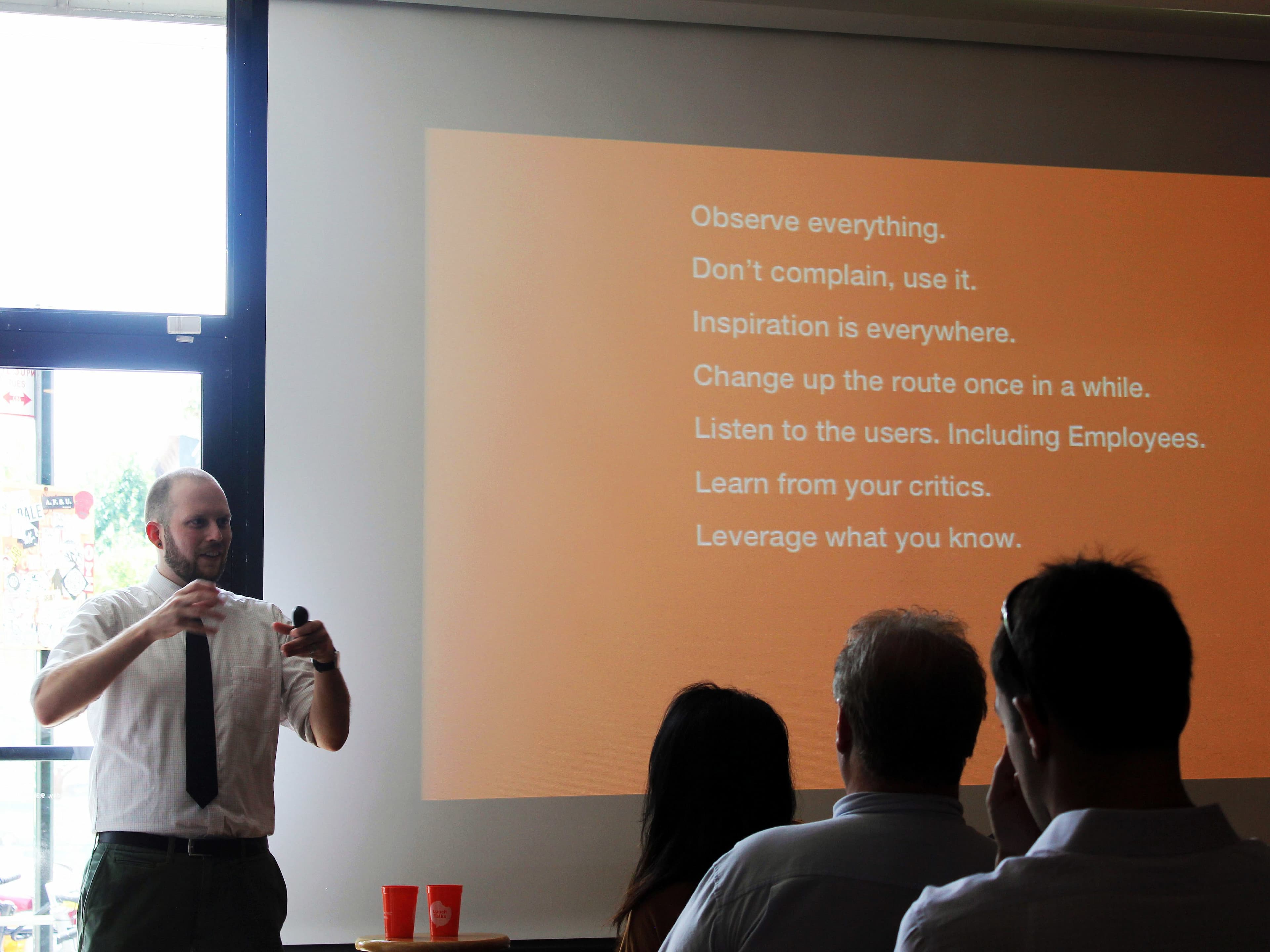 A man in a white shirt and tie is giving a presentation in a room with several people seated. Behind him, an orange slide with text is projected on the screen that includes tips such as "Observe everything" and "Inspiration is everywhere.