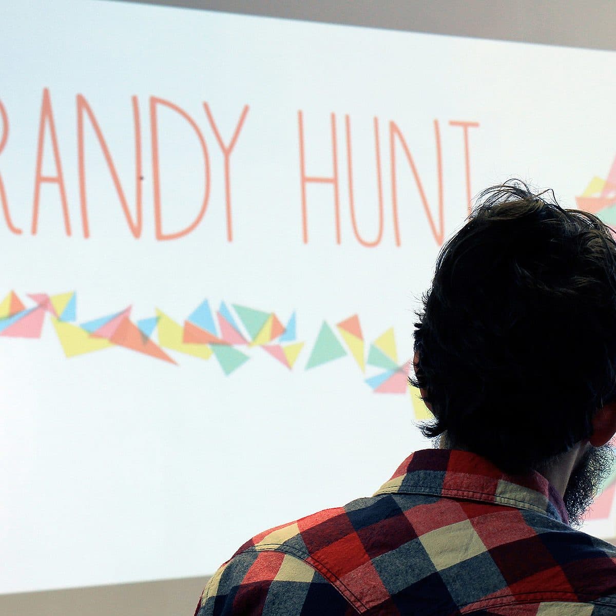 A person with a beard, wearing a checkered shirt, is facing a screen with the name "RANDY HUNT" displayed in large letters. The text is adorned with colorful geometric shapes.