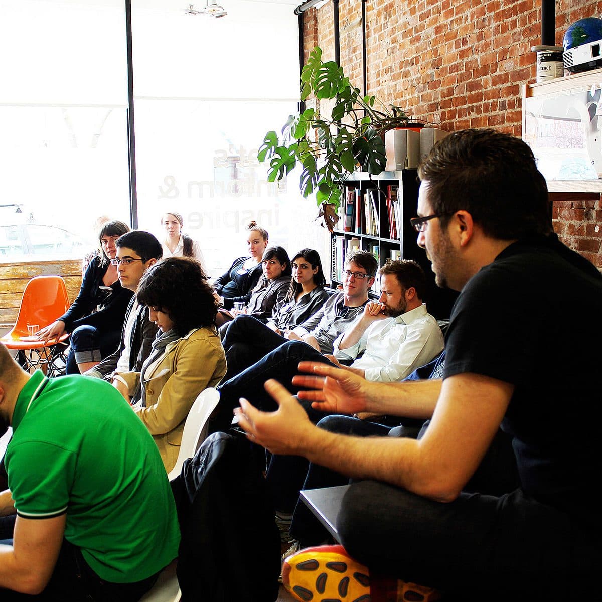 A group of people sit in a casual meeting room with brick walls. They are attentively listening to a man in a black shirt who is speaking and gesturing. There is a large window in the background, some orange chairs, and plants, creating a relaxed atmosphere.