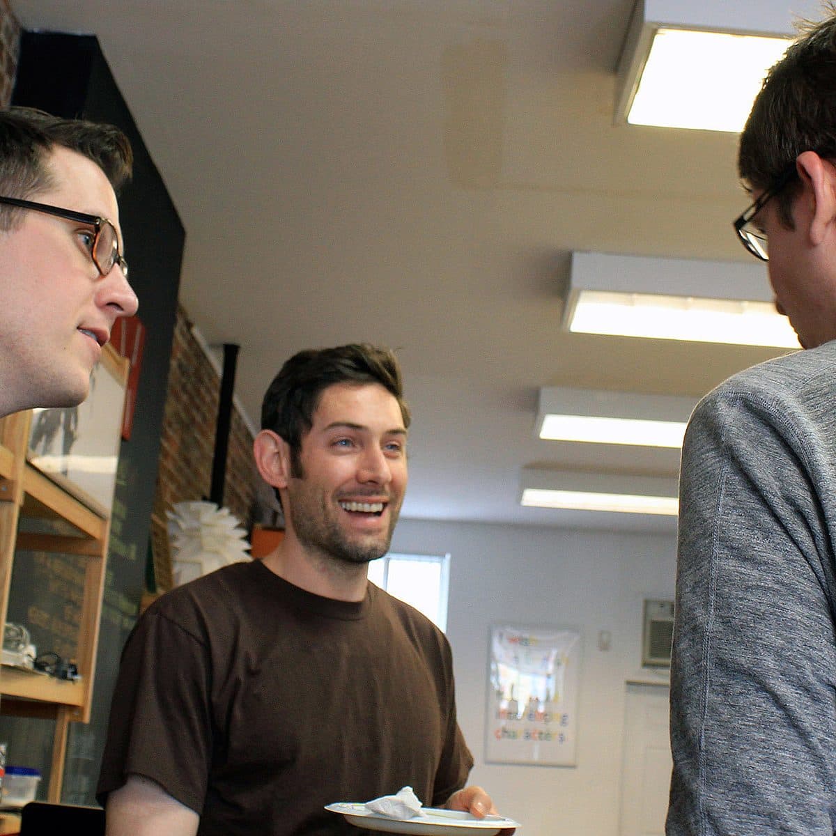 Three people are gathered indoors, engaged in conversation. One person in the middle is smiling and holding a small white plate. The environment suggests a casual setting with wooden shelves and fluorescent lighting visible in the background.