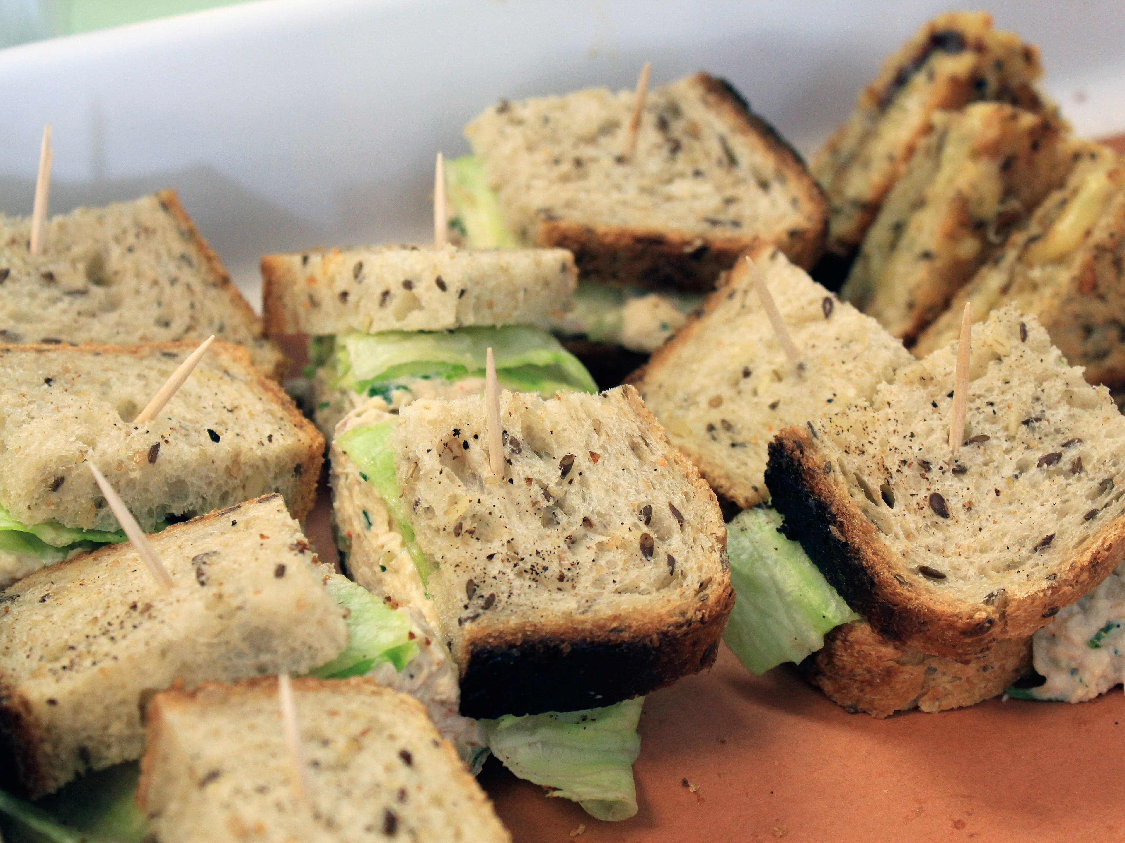 A tray filled with small sandwich squares made with whole grain bread. Each sandwich is held together with a toothpick and contains lettuce and a creamy filling. The sandwiches appear fresh and are arranged neatly in the tray.