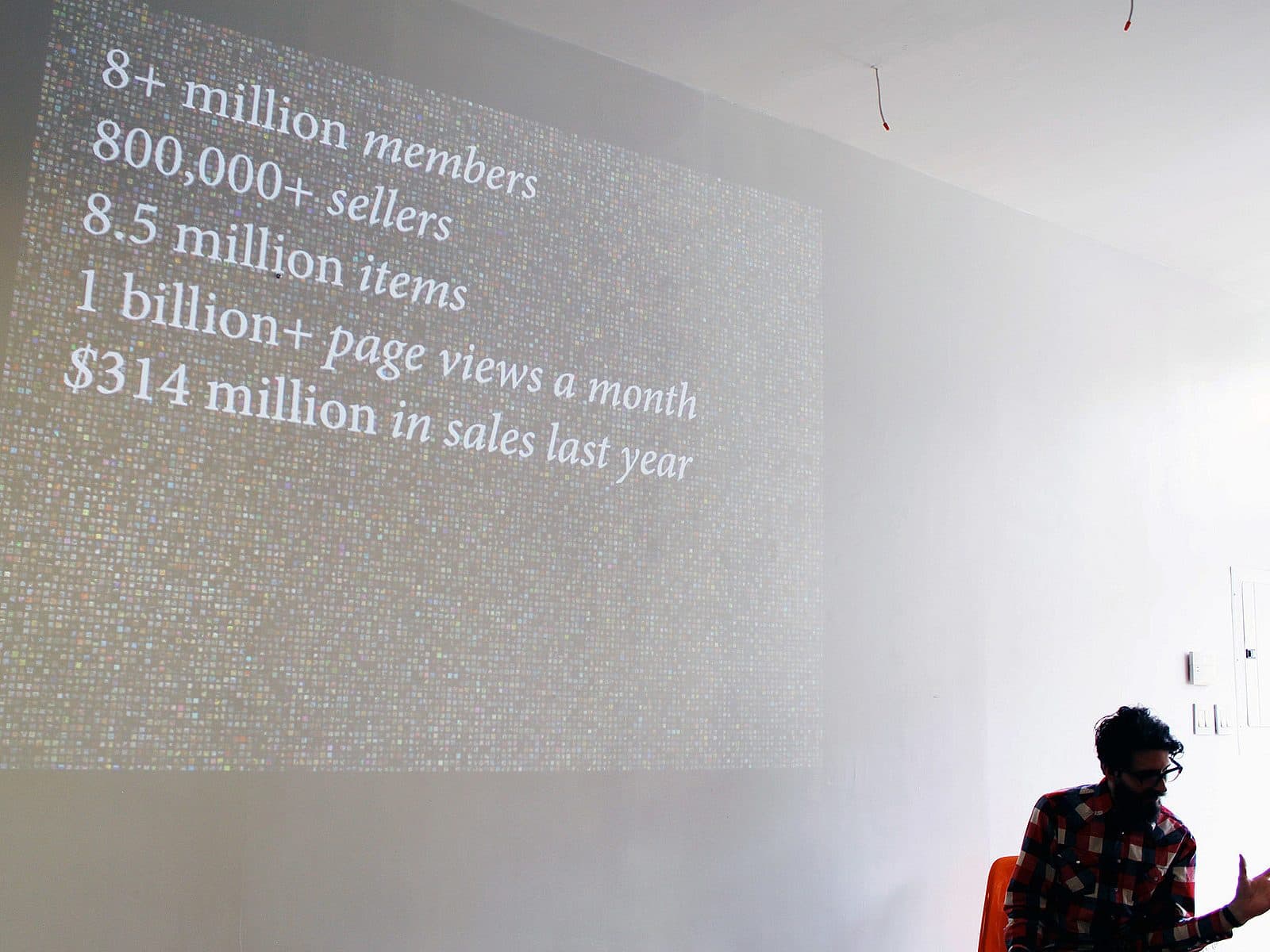 A person with glasses and a beard, seated, gives a presentation in a room lit by natural light. The screen displays text: "8+ million members, 800,000+ sellers, 8.5 million items, 1 billion+ page views a month, $314 million in sales last year.