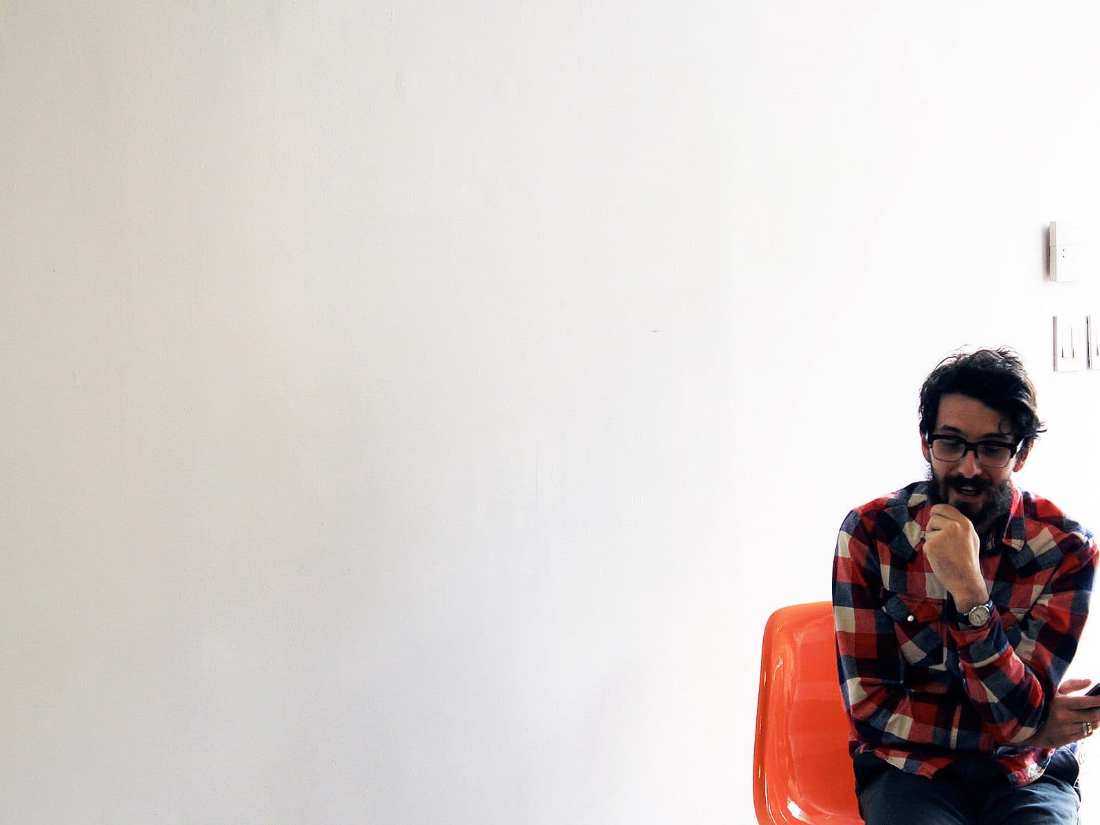 A man with glasses and a beard sits on an orange chair against a white wall. He is wearing a red and white plaid shirt and is holding a smartphone in his right hand while resting his left hand near his mouth. The room appears minimalistic.