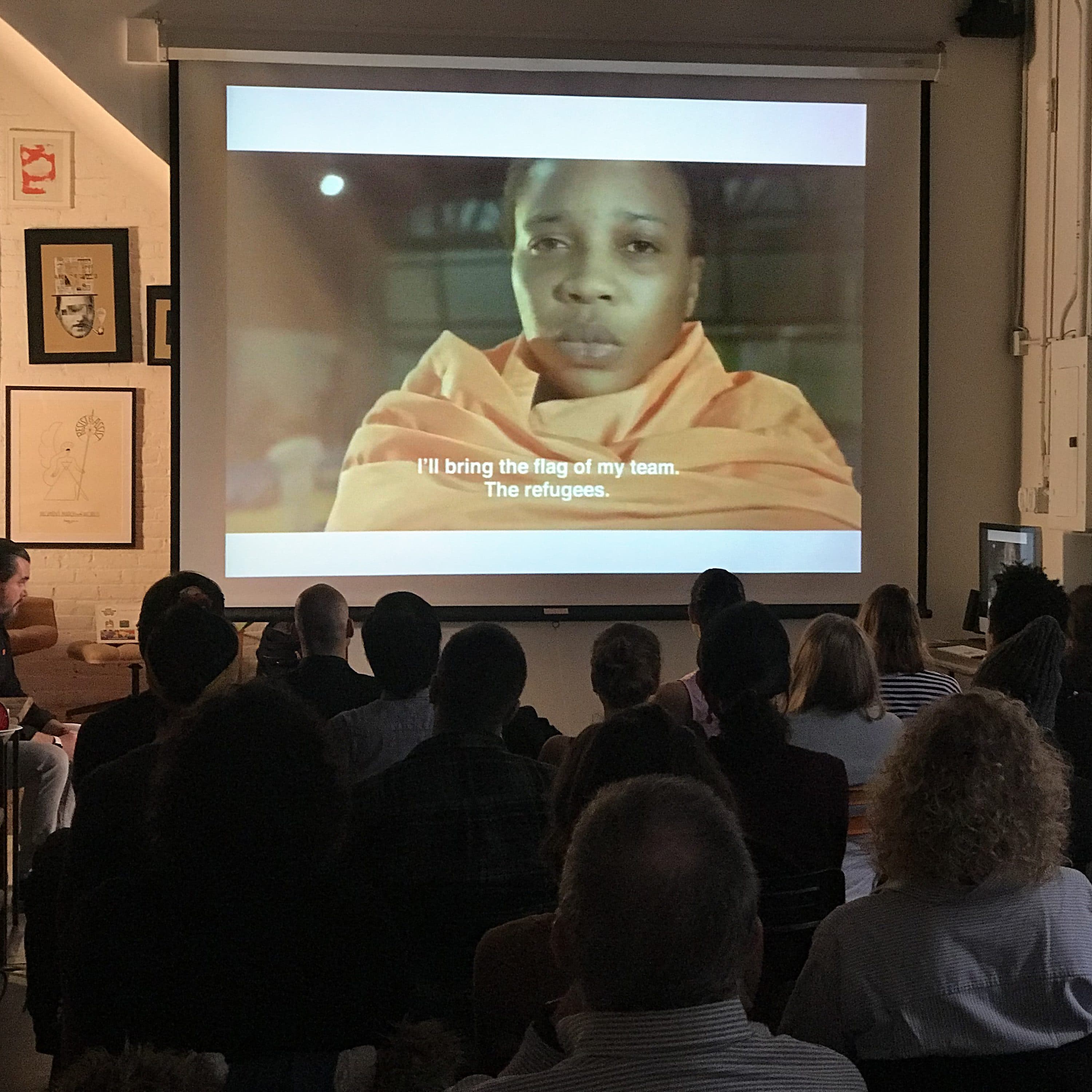 A group of people seated in a dimly lit room watches a film projected on a screen. The film shows a person wrapped in an orange blanket with the subtitle, "I'll bring the flag of my team. The refugees." Art and posters are displayed on the room's walls.