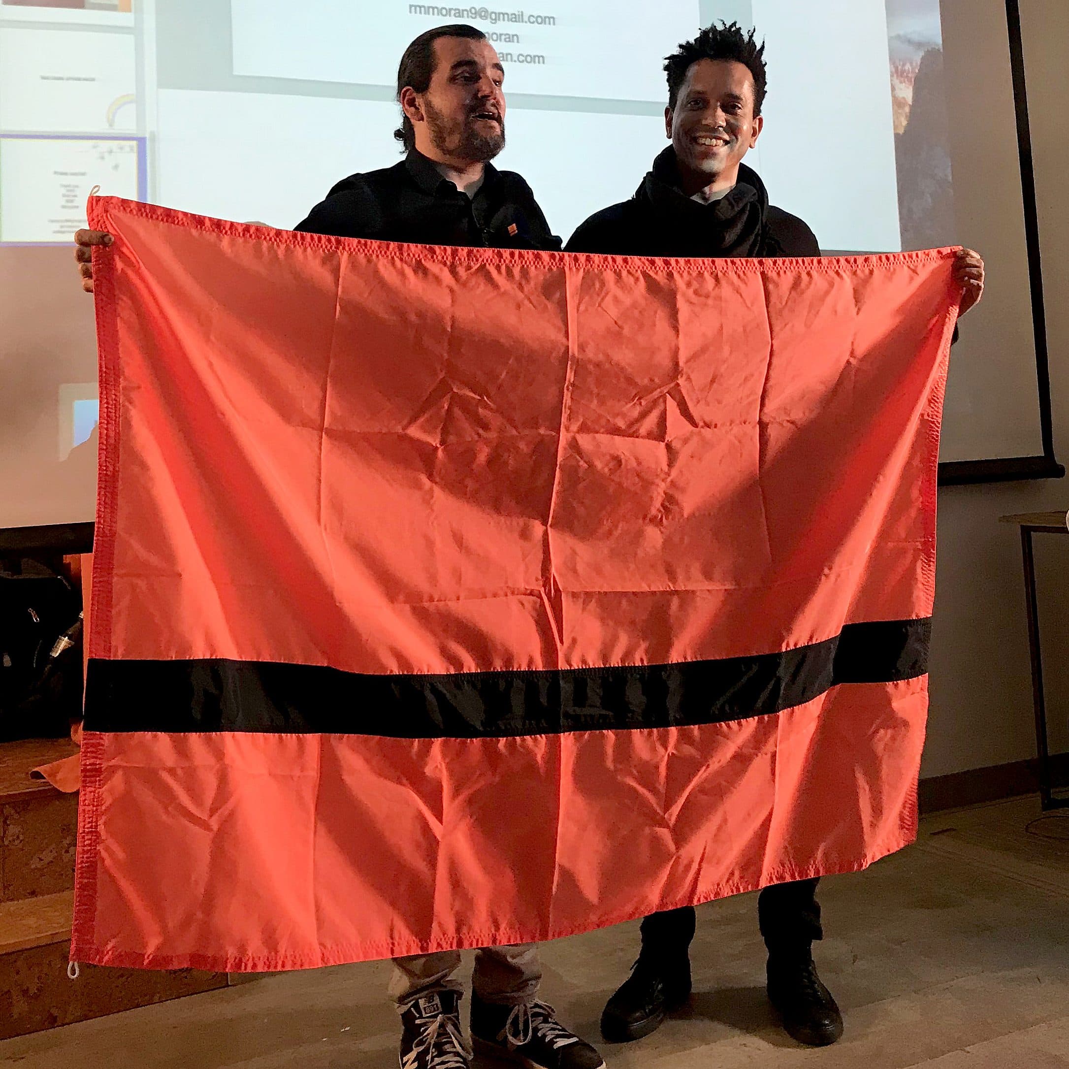 Two people stand indoors holding a large red flag with a thick black horizontal stripe in the center. They are smiling, and a projector screen displaying an email address is visible behind them. Both are casually dressed, one in a black hoodie and the other in a black shirt.