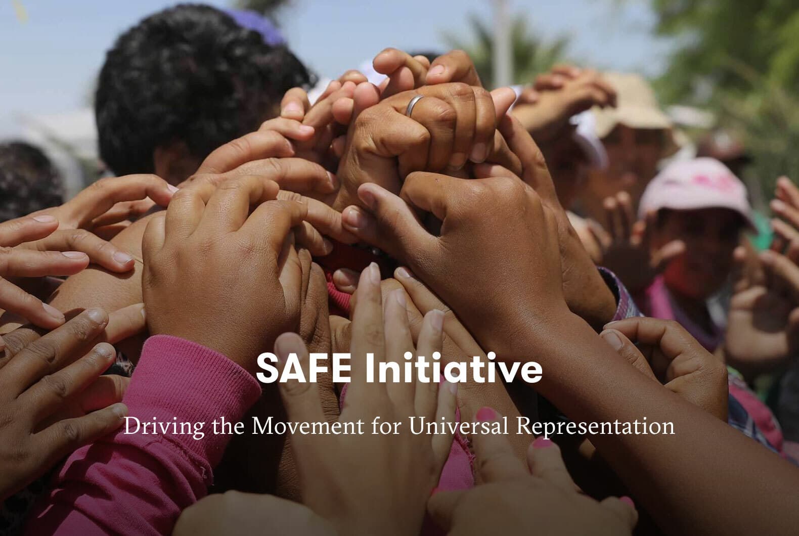 A group of diverse hands clasping together in solidarity, highlighting the SAFE Initiative with the slogan "Driving the Movement for Universal Representation" written over the image. The background is blurred with people engaging in the movement.