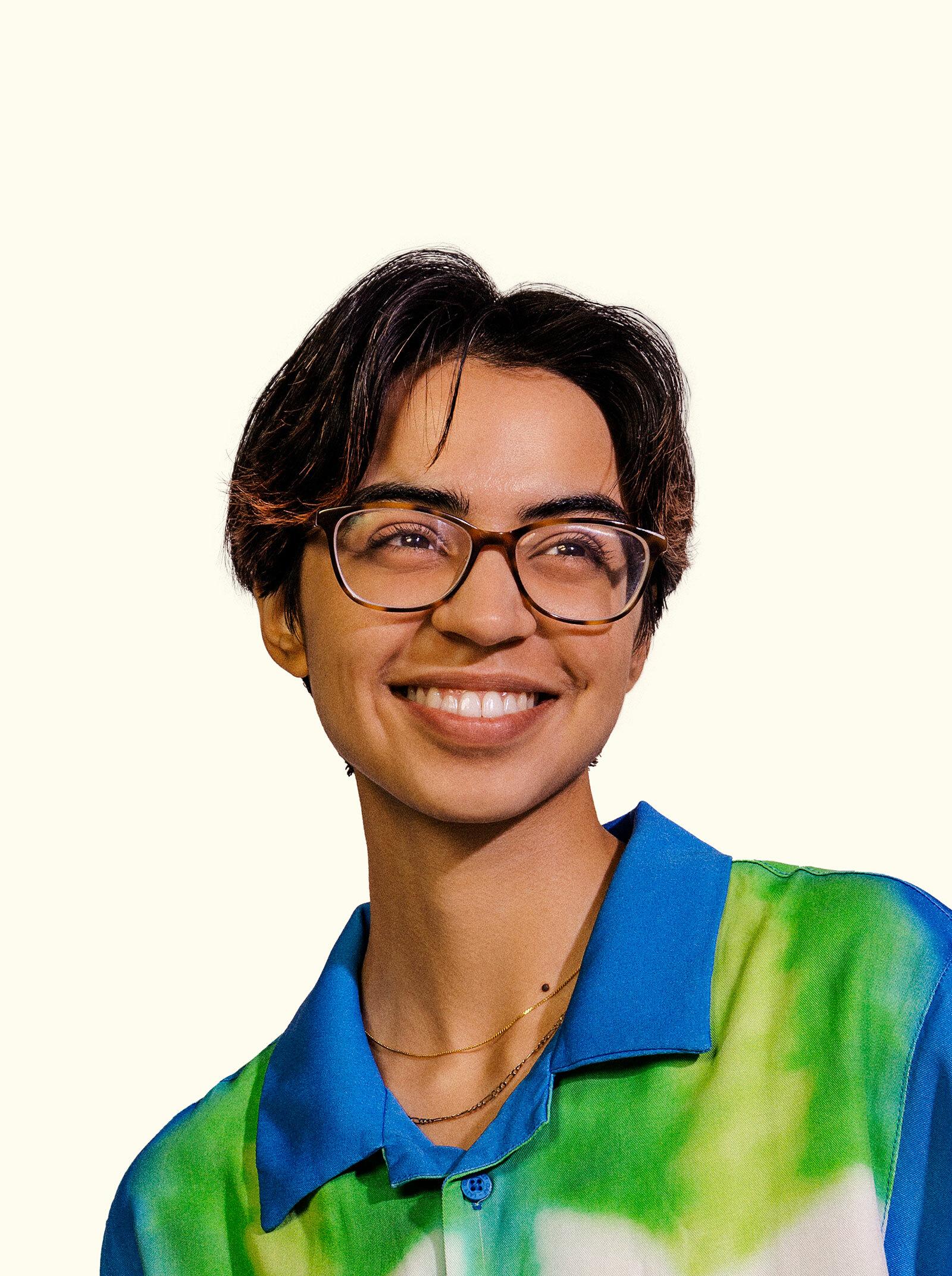 A person with short dark hair, wearing glasses and a brightly colored blue and green shirt, smiles warmly. They have a light complexion and are looking slightly off to the side. The background is a plain light color.