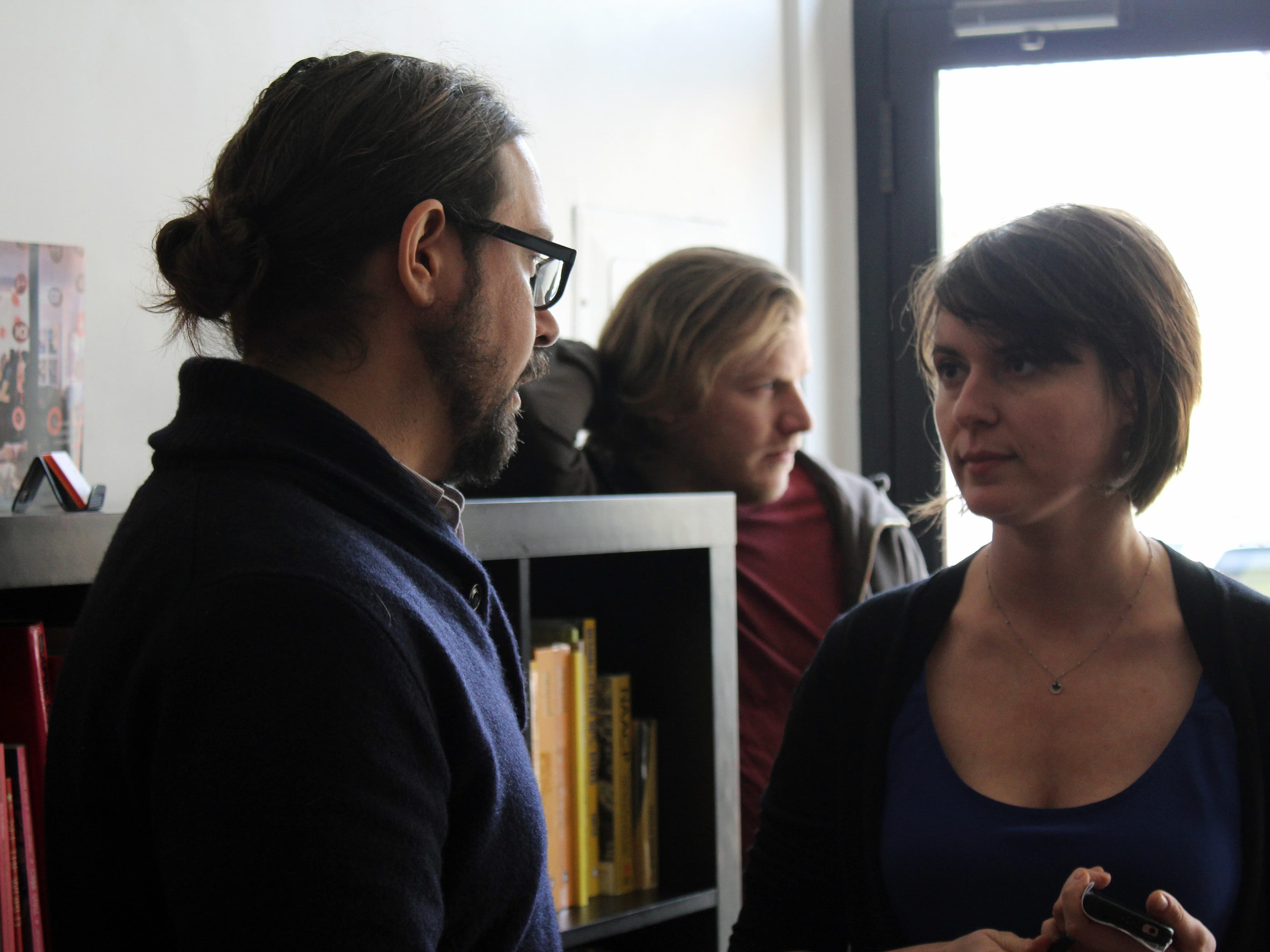 A man with glasses and a ponytail is speaking to a woman with short hair who is holding a phone. Another man with blond hair stands in the background, facing away. They are indoors near a window, with a bookshelf filled with books next to them.