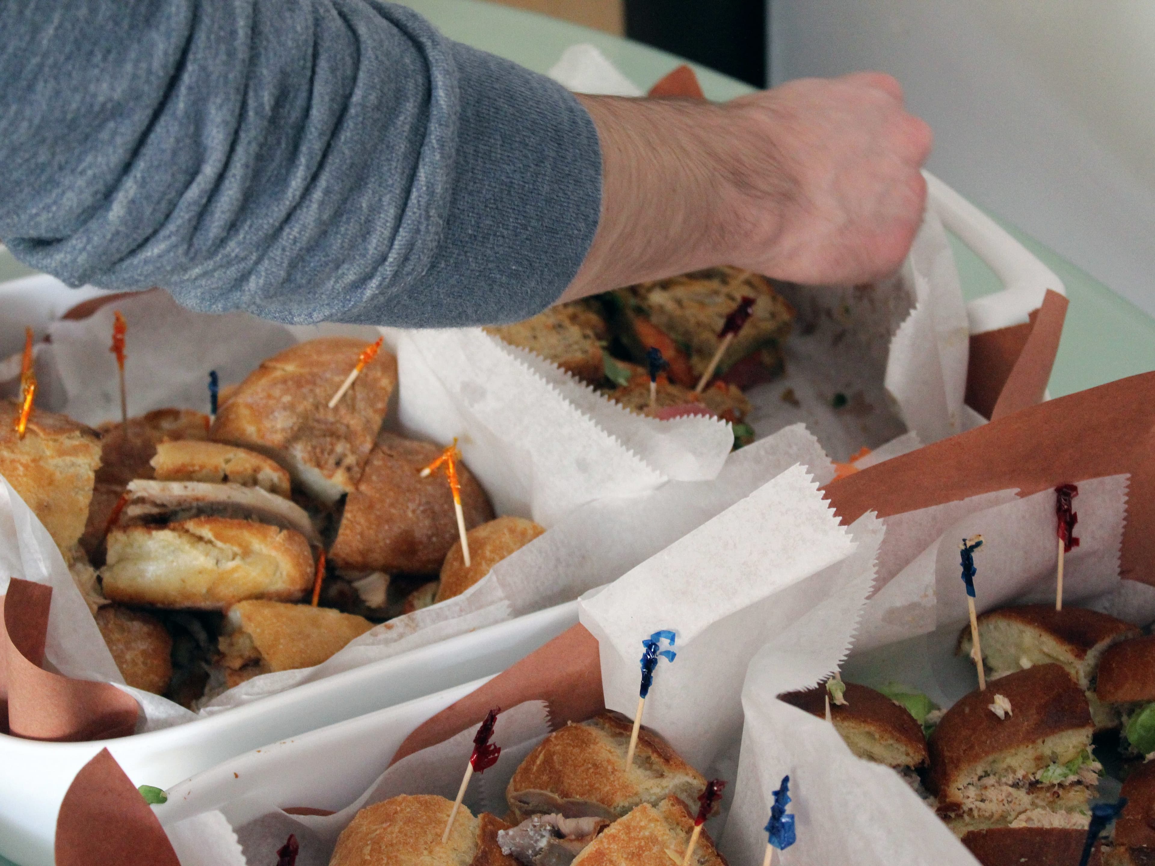 A person reaches for a sandwich from a platter filled with various sandwich pieces. Each piece is secured with a colorful toothpick. The sandwiches are displayed on white trays lined with parchment paper. The person is wearing a gray long-sleeve shirt.
