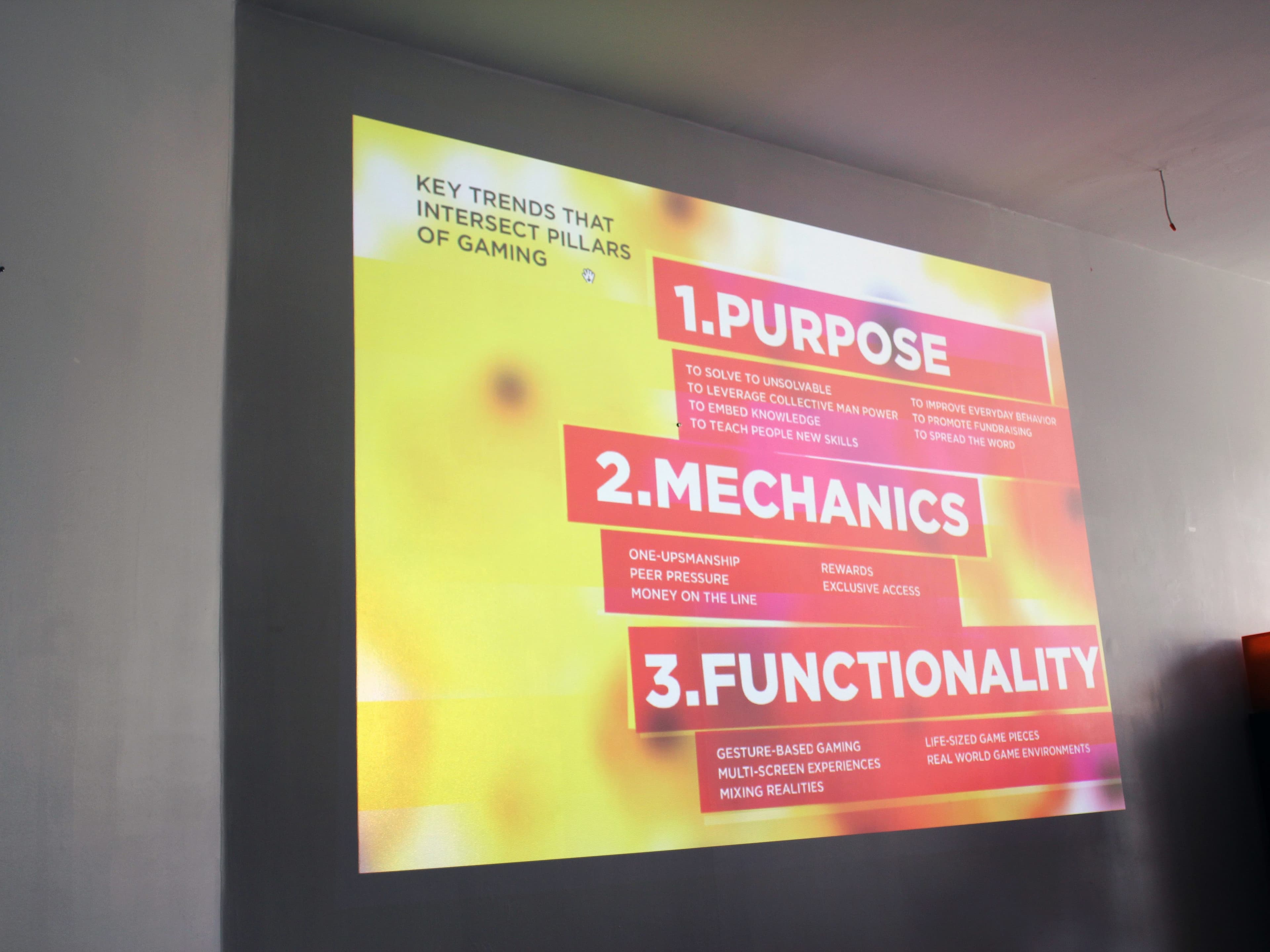 A brightly lit projector screen displays three key trends intersecting gaming pillars: 1. Purpose, 2. Mechanics, and 3. Functionality. Each trend is elaborated with bullet points in white text against red and orange highlighted areas on a gradient yellow and orange background.