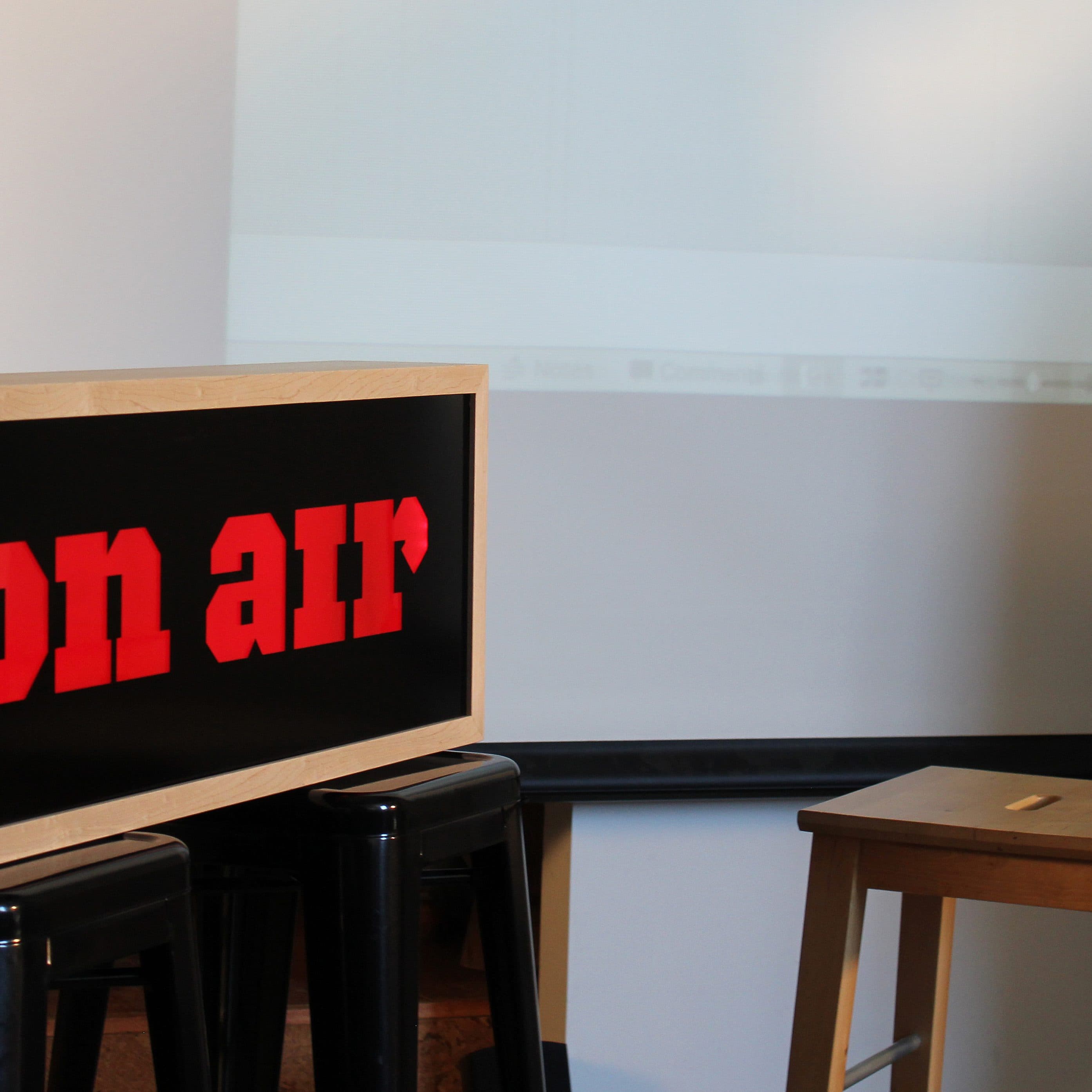 A wooden "on air" sign with red lettering sits on a black stool next to a plain wooden stool. The background includes part of a projection screen and a white wall, suggesting a studio or recording setup.