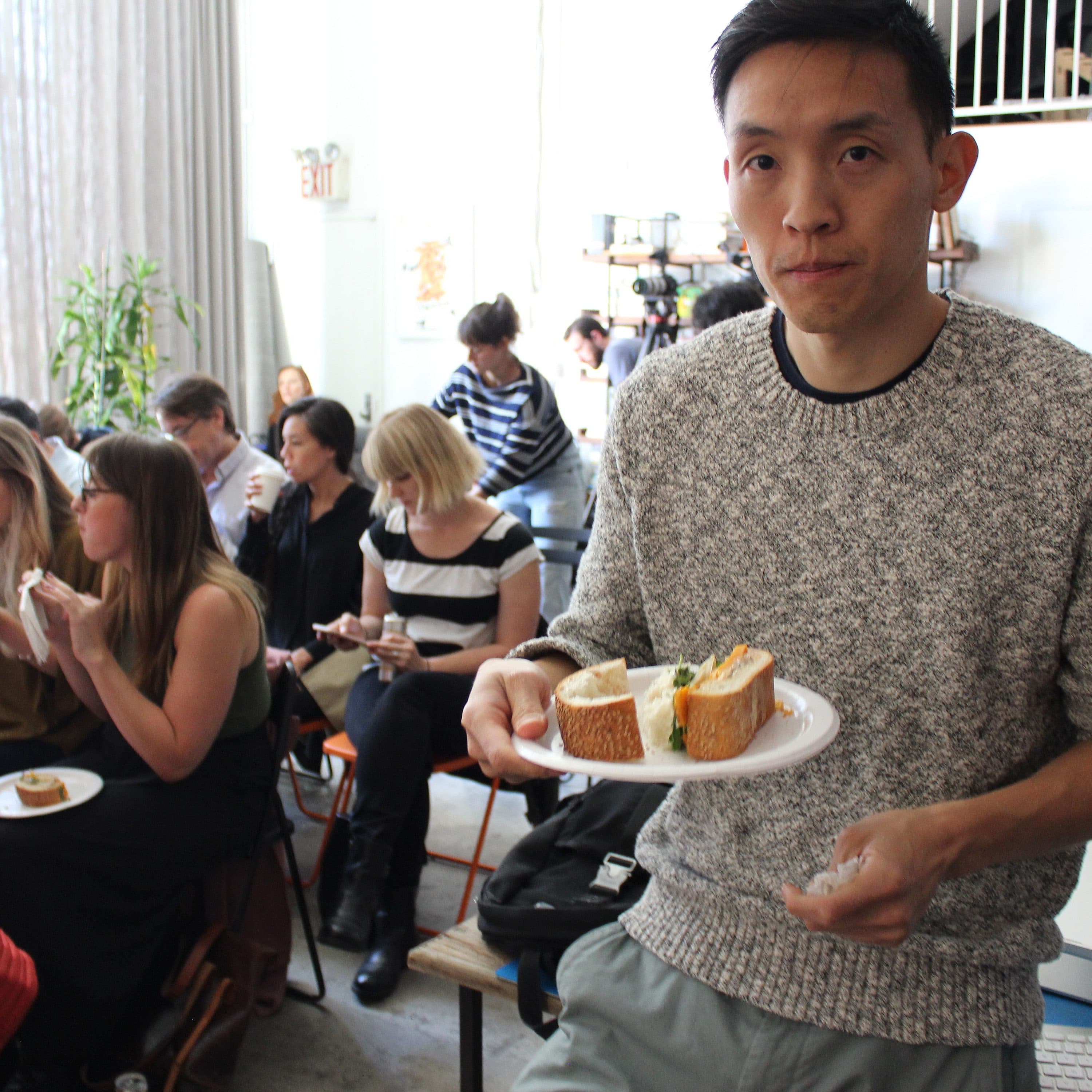 A man wearing a gray sweater holds a plate with a sandwich, standing in a room where multiple people sit and eat sandwiches. The room has a modern design with plants, large windows, and a loft area.