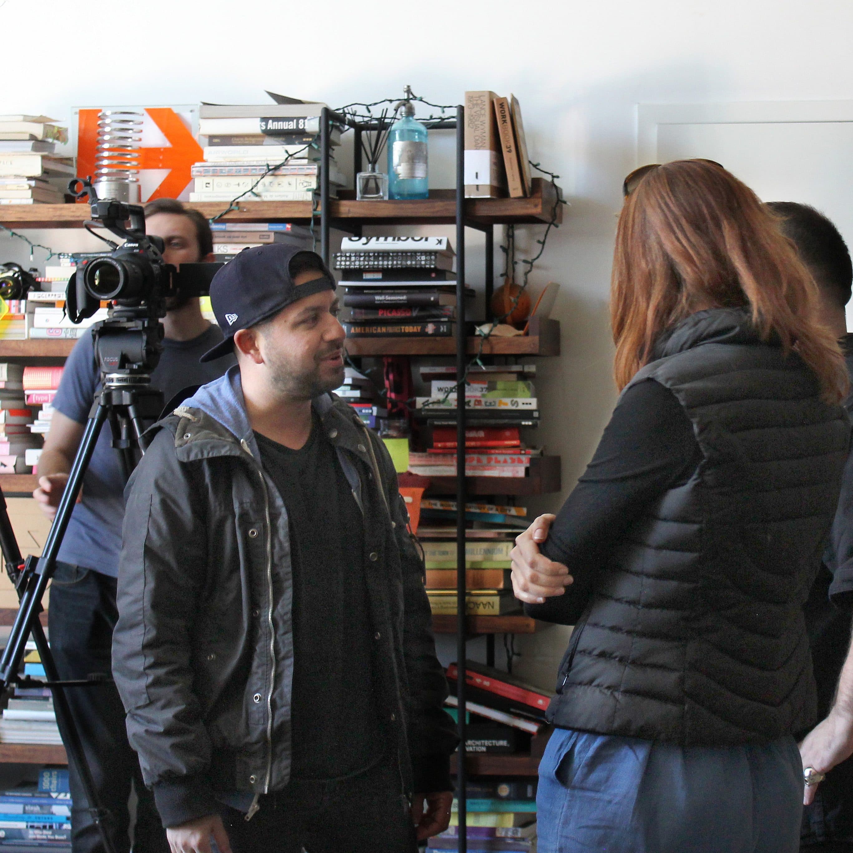 People standing and talking in a room with bookshelves and video cameras on tripods in the background. One man is wearing a backward cap and a jacket, while the woman has long hair and is wearing a puffer vest. There are books and various objects on the shelves.