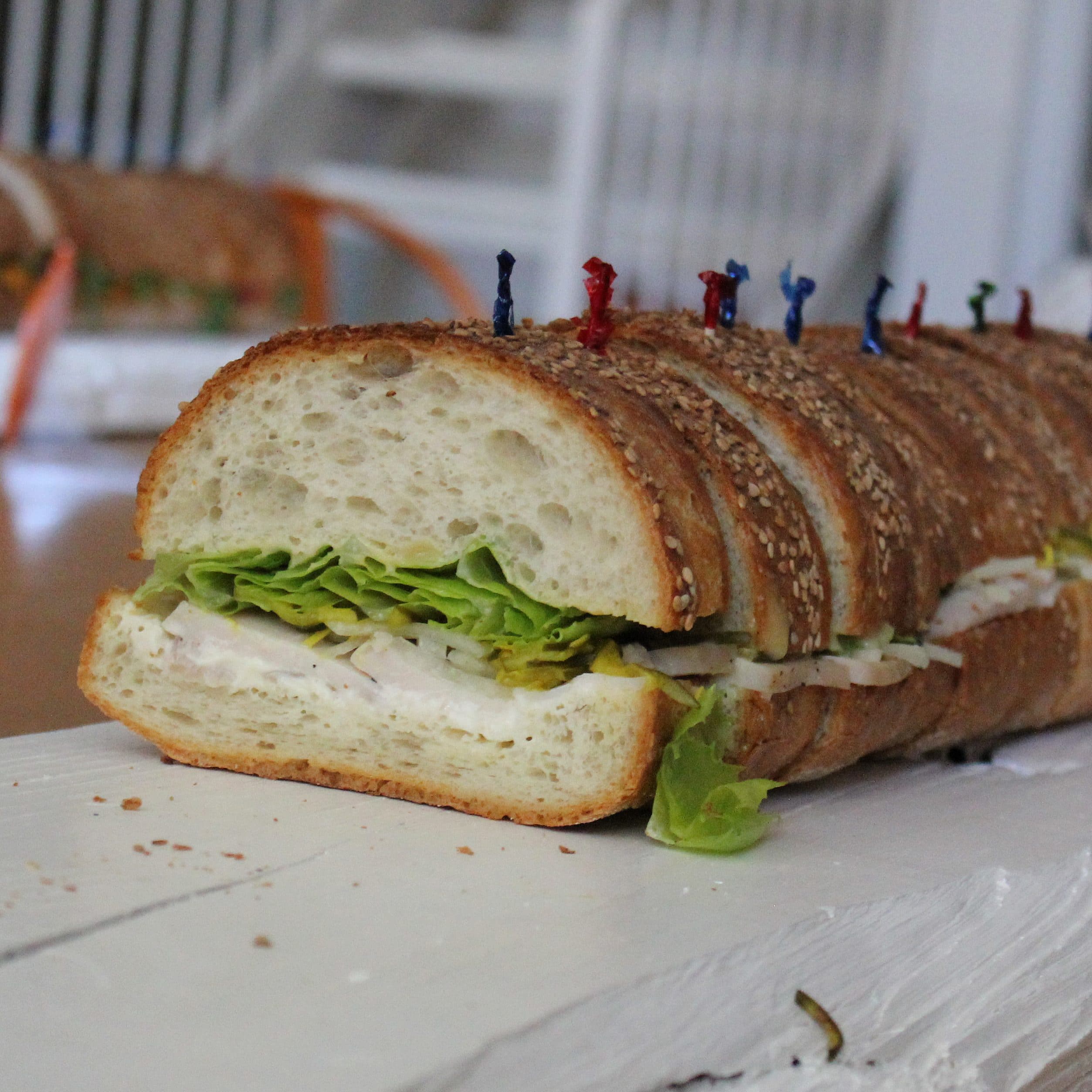 A long sandwich on a white tray is filled with lettuce, cheese, and meats. The sandwich is topped with colorful toothpicks to hold it together. In the background, more sandwiches are partially visible.